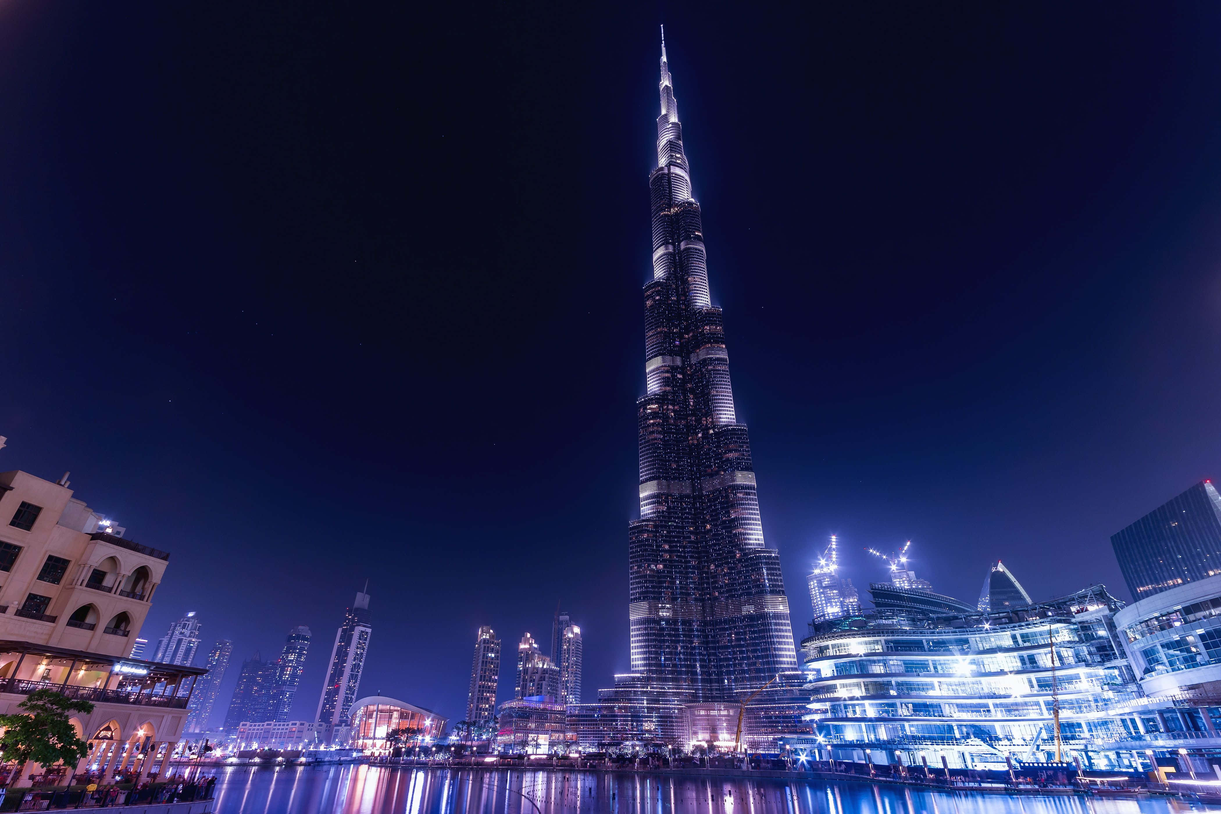 Dubai 桌布 壁紙 — 哈利法塔在夜晚杜拜的天際線上高聳入雲，其發光的尖頂倒映在下方水面上。