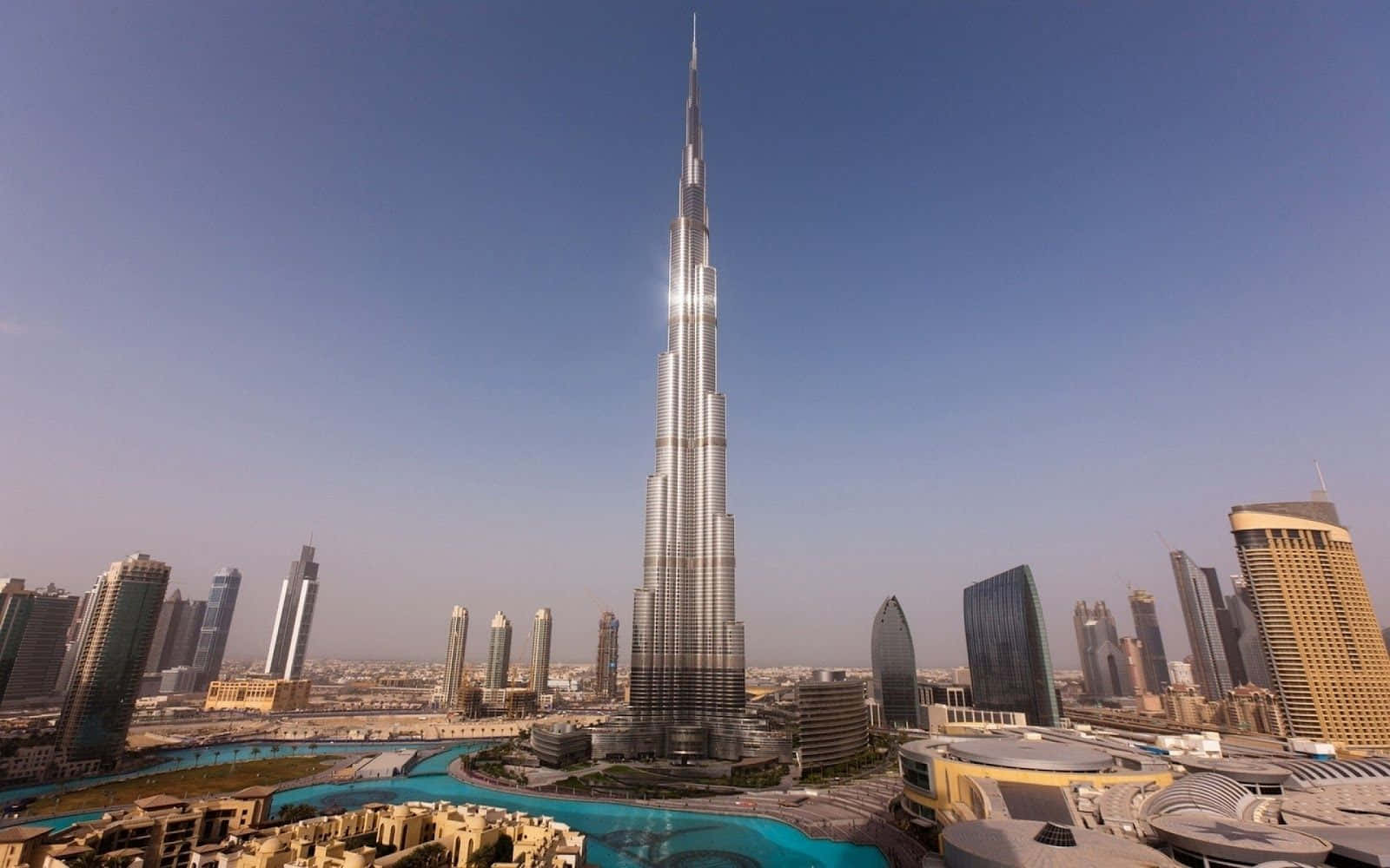 Burj Khalifa 桌布 壁紙 — 哈利法塔聳立在杜拜天際線上，其閃閃發光的尖頂刺穿清澈的藍天，下方是現代玻璃摩天大樓和藍綠色水景。