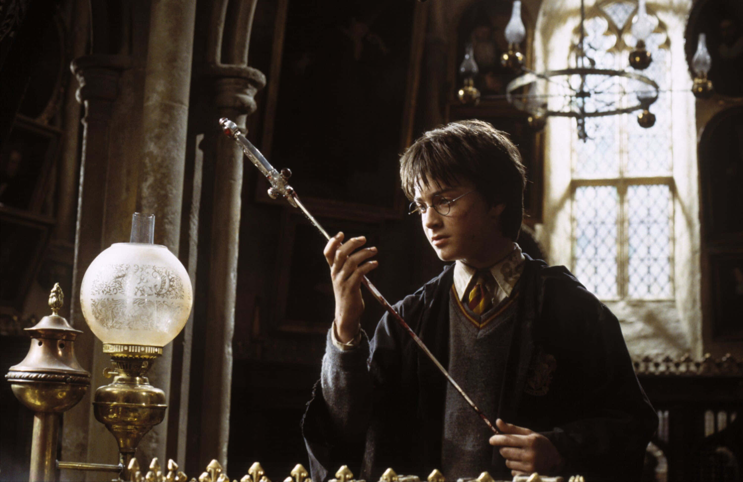 The Chamber Of Secrets 桌布 壁紙 — 哈利波特在一個光線昏暗、裝飾華麗的中世紀風格教室中，專注地看著他的魔杖，教室裡有石柱和彩繪玻璃窗。