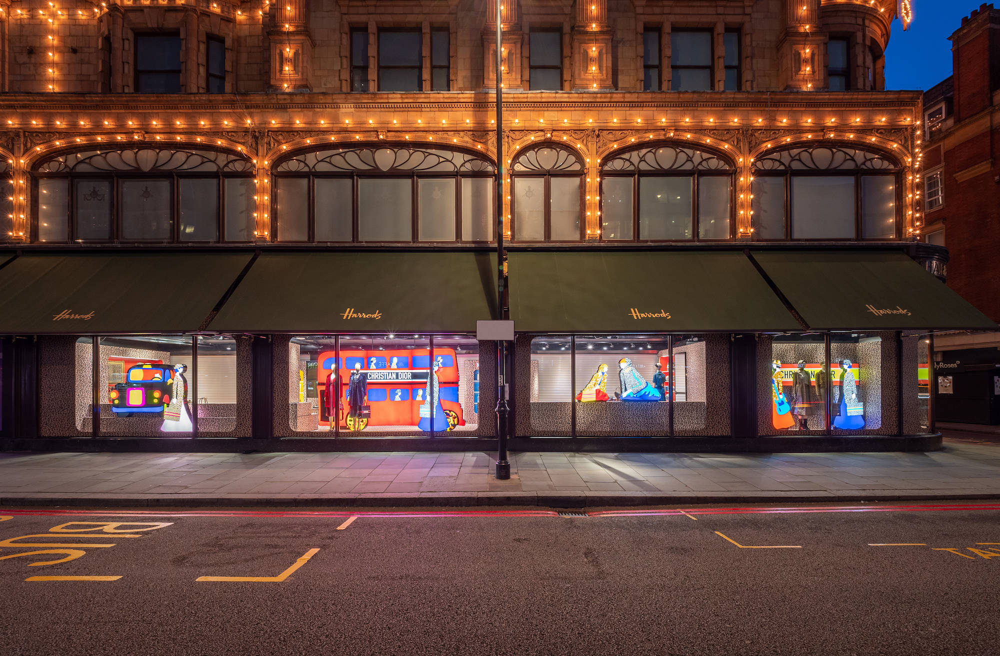 哈洛德夜間精品店 壁紙 — Harrods 百貨公司外牆在黃昏時分，溫暖的串燈在色彩繽紛的櫥窗展示上方閃爍。