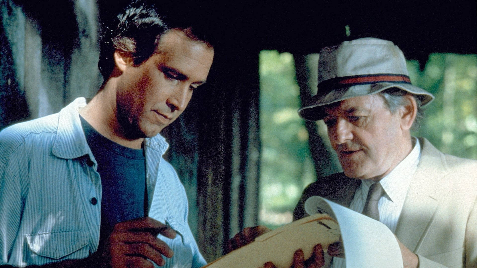 哈爾·霍布魯克在《Fletch Lives》（1989）經典畫面 壁紙 — 兩個男人在森林小屋中一起仔細檢查一張捲起的藍圖。