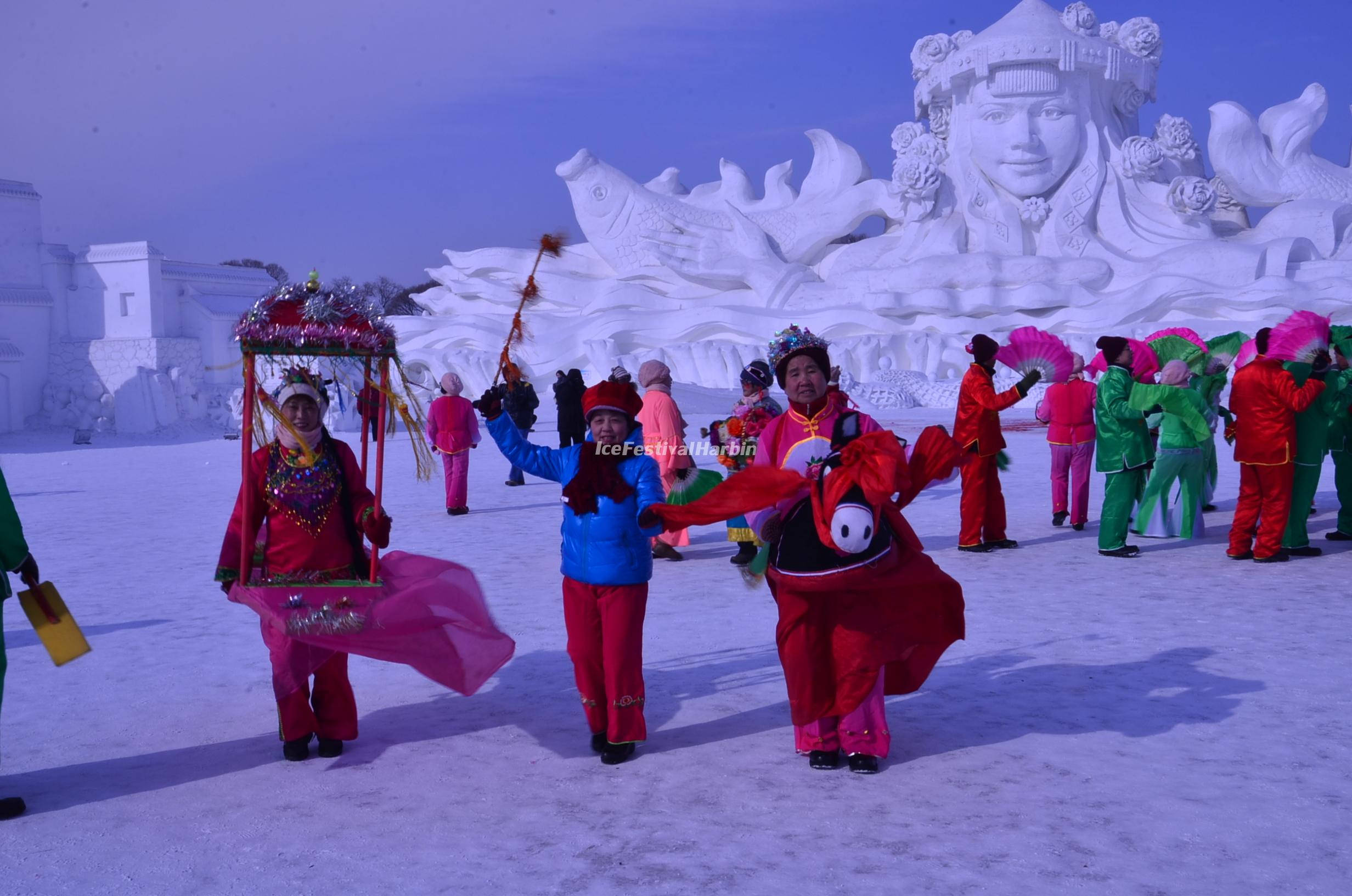 哈爾濱冰雪大世界壯觀冰雕展 壁紙 — 色彩繽紛的表演者在雪地上歡快地跳舞，背景是一座巨大而精緻的雪雕。