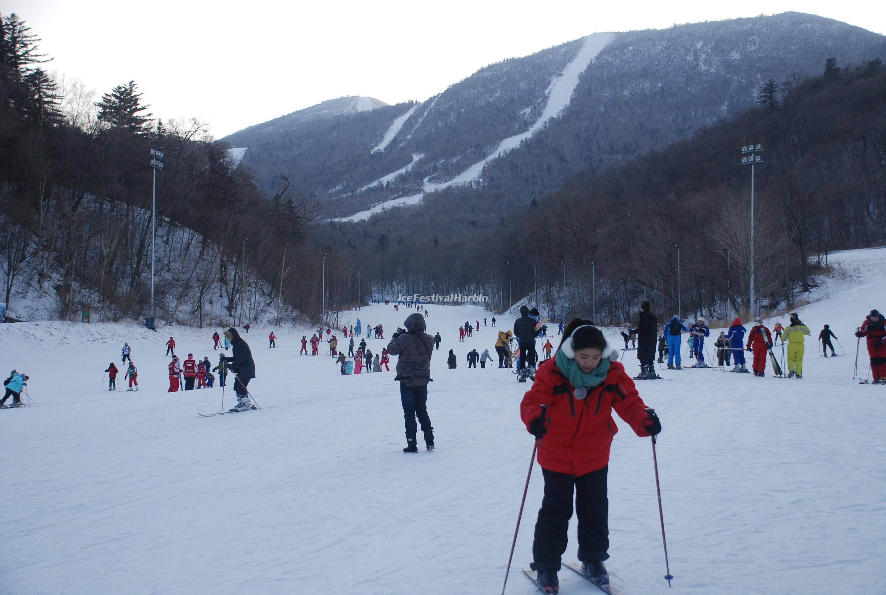 哈爾濱城市日落全景 壁紙 — 穿著色彩繽紛裝備的滑雪者，在蒼白的冬日天空下滑下白雪覆蓋的山坡。