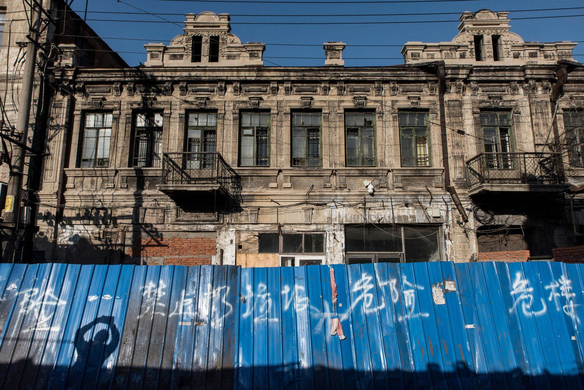哈爾濱舊建築 壁紙 — 陽光照射下破舊的華麗建築，前景有藍色波紋柵欄投下長長的影子。