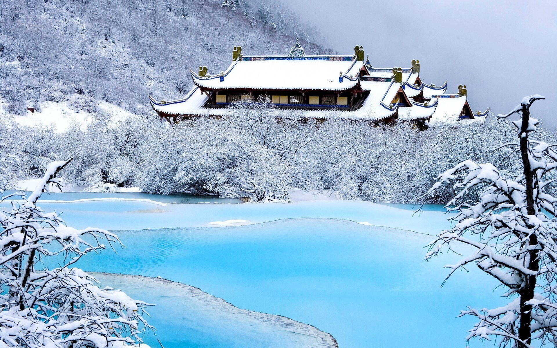 哈爾濱雪山覆雪屋舍 壁紙 — 一座被雪覆蓋的傳統寺廟坐落在充滿活力的碧綠湖泊之上，周圍環繞著霜凍的樹木和霧濛濛的山脈。