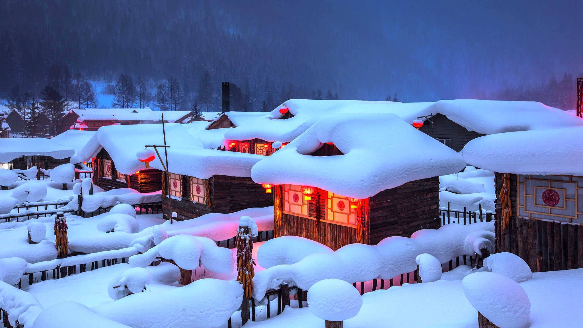 哈爾濱雪村 壁紙 — 白雪覆蓋的木屋在深藍色的冬夜天空下溫暖地發光。