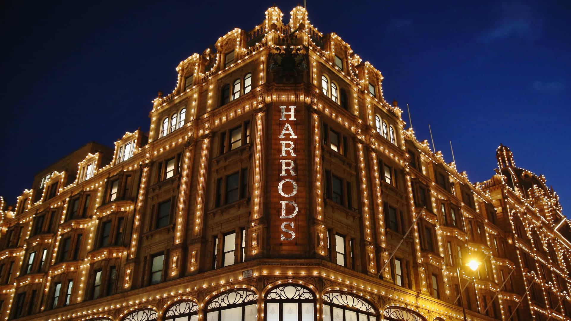 哈羅德百貨深藍天空 壁紙 — Harrods 百貨公司在深藍色的夜空下溫暖地發光，其華麗的外牆裝飾著閃爍的燈串。