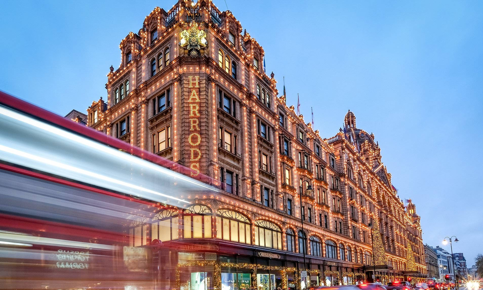 哈羅德藍天 壁紙 — Harrods 百貨公司在暮色天空下溫暖地發光，一輛紅色雙層巴士以動態模糊效果疾馳而過。