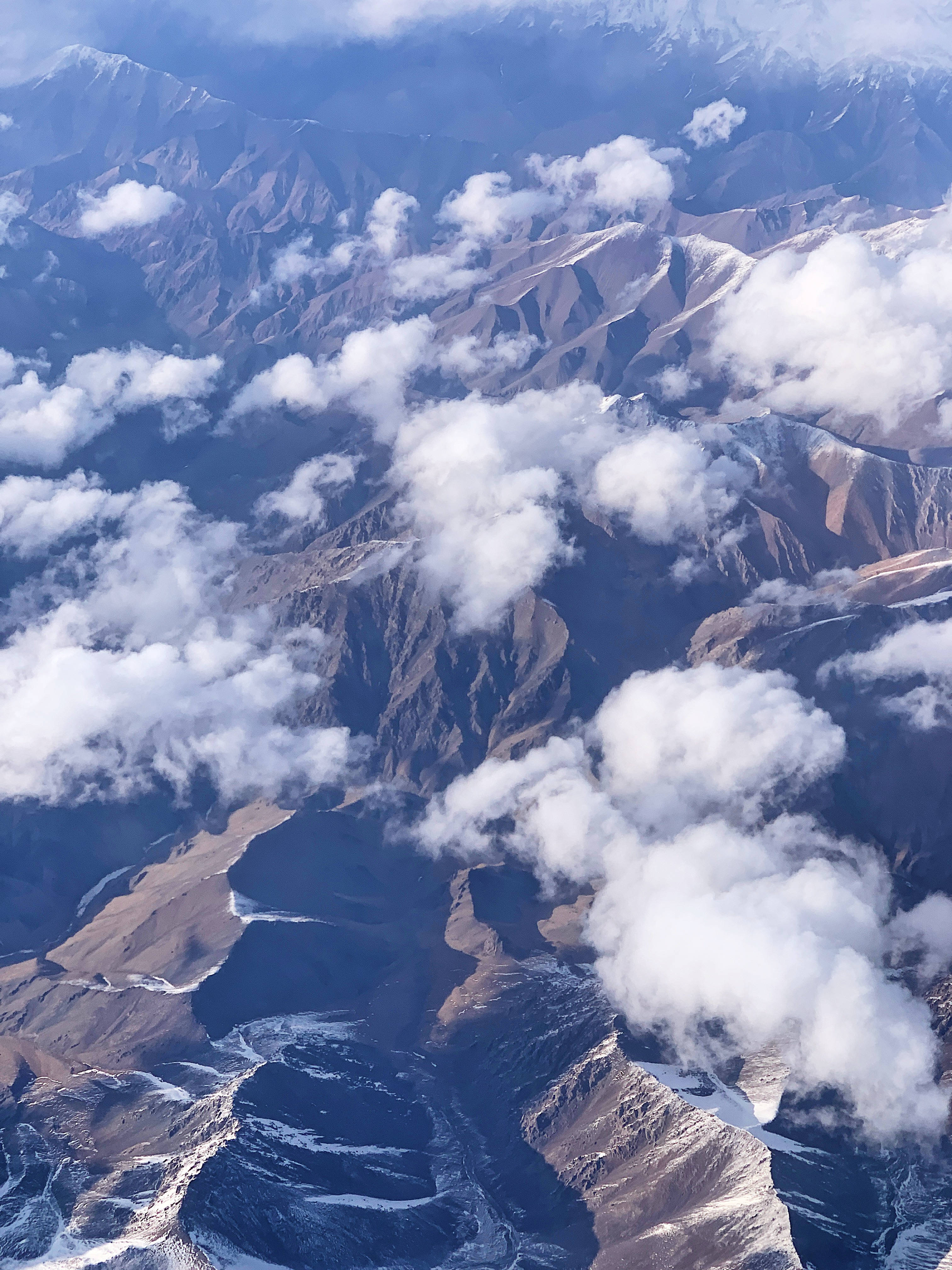 哈薩克斯坦山脈空中視角 壁紙 — 崎嶇山脈的鳥瞰圖，山頂積雪，蓬鬆的白雲在山谷間飄移。