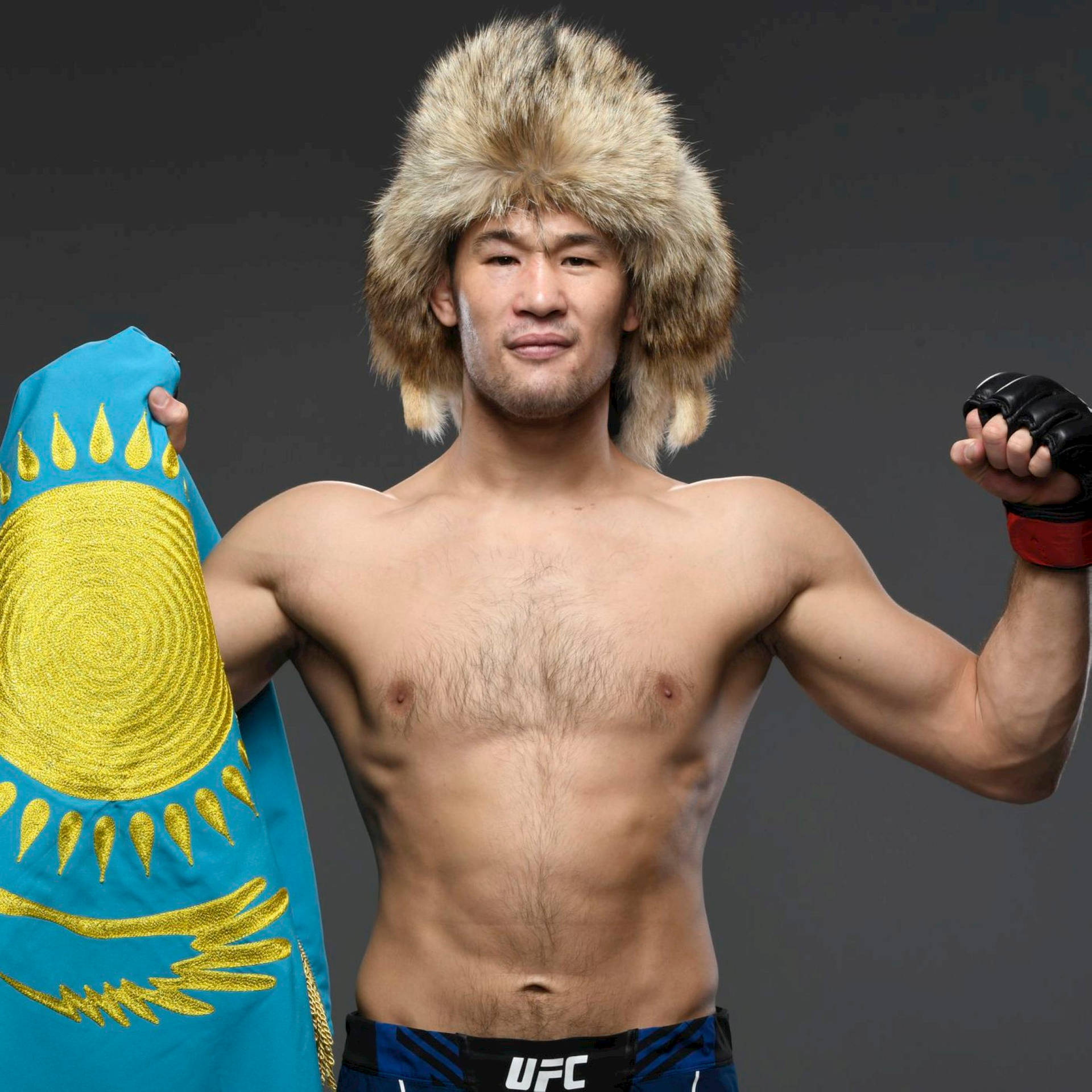 一位赤裸上身的哈薩克戰士戴著毛帽，手持國旗並與UFC腰帶一起擺姿勢。