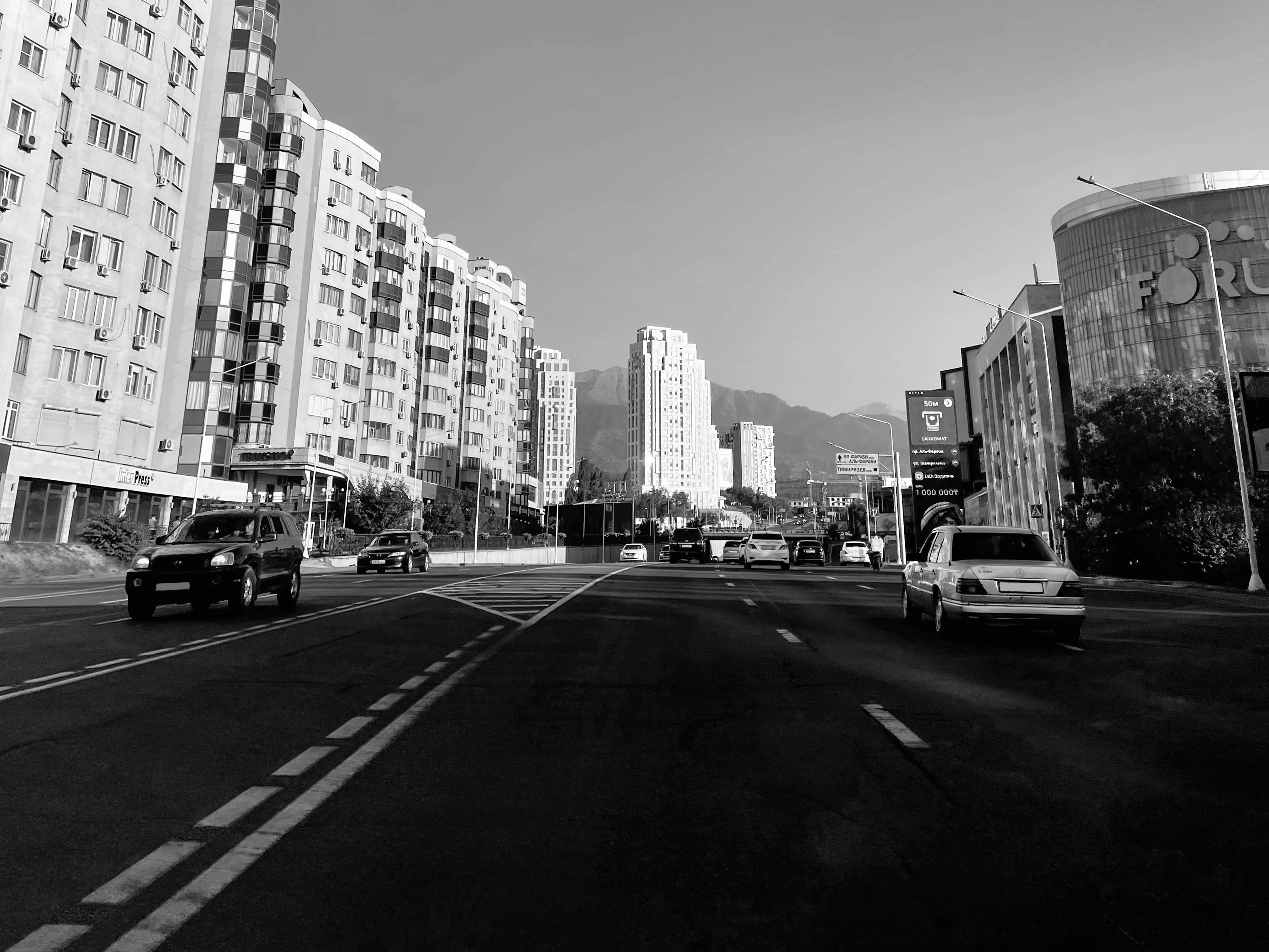 哈薩克斯坦黑白攝影 壁紙 — 黑白城市街景，汽車駛過高樓大廈和遠山，天空晴朗。