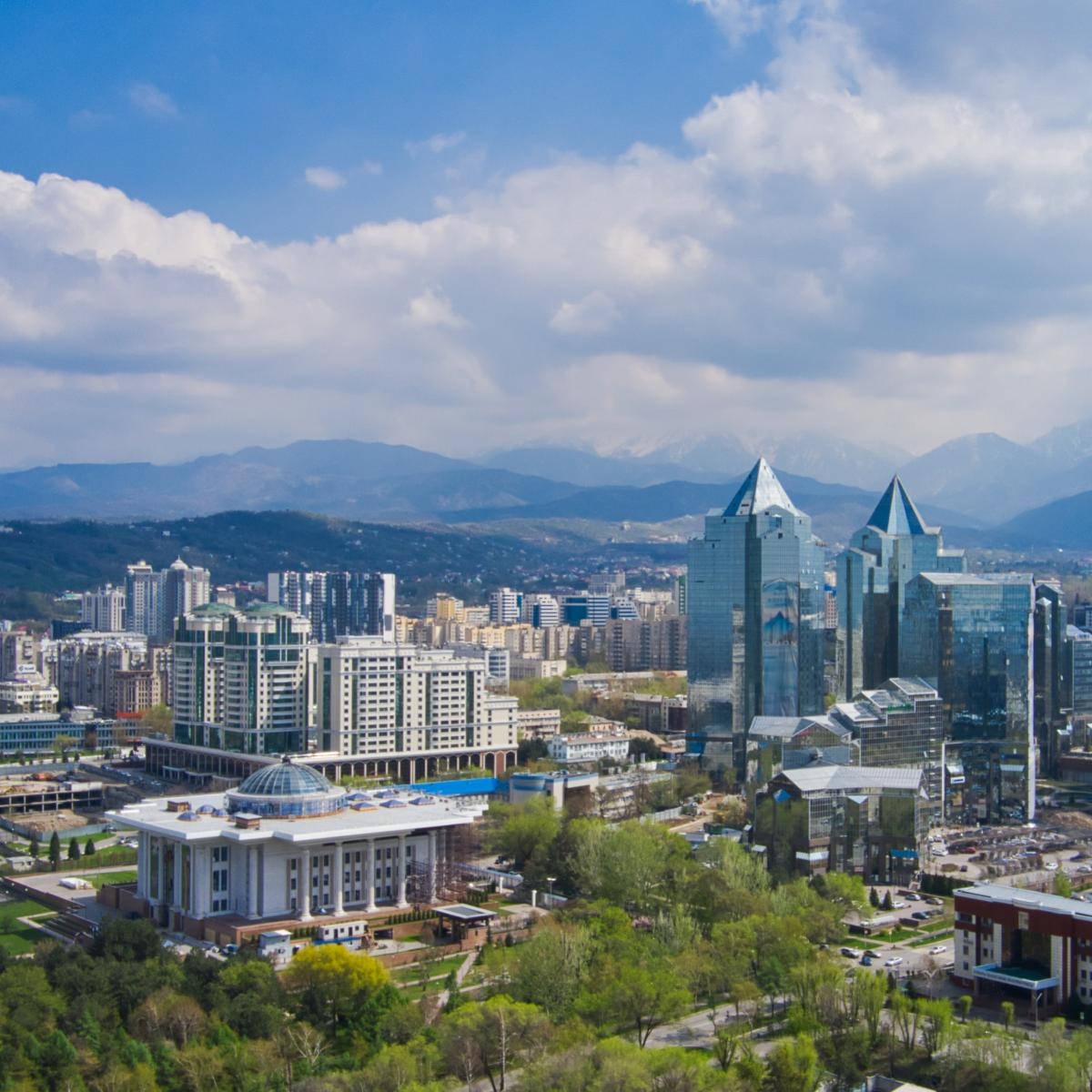 哈薩克阿拉木圖城市風景 壁紙 — 現代城市景觀的鳥瞰圖，玻璃摩天大樓和綠色公園在明亮的藍天下，有蓬鬆的雲朵和遠處的雪山。