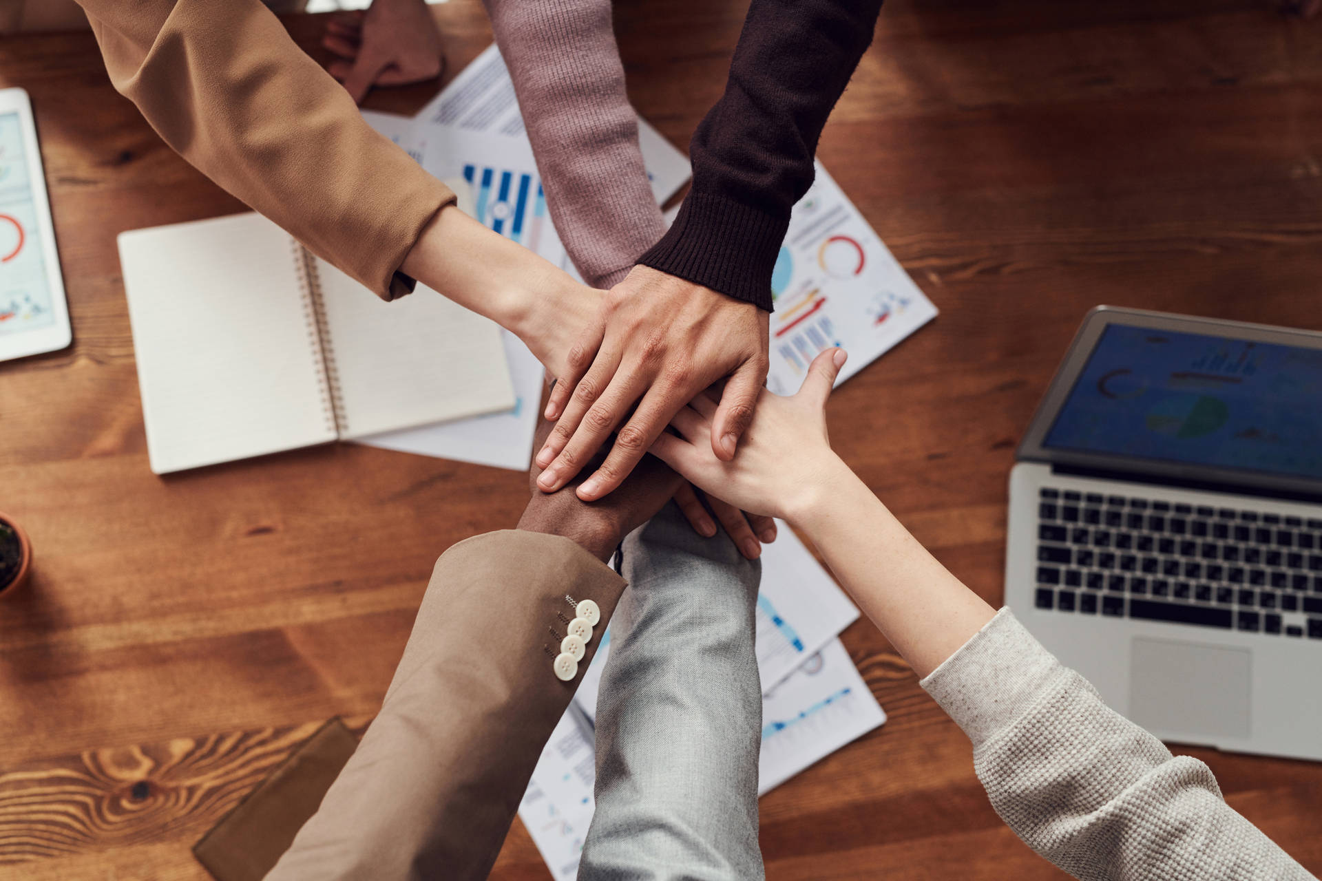 在木桌上，不同人士的手疊放在一起，進行商業會議，周圍有圖表和手提電腦。
