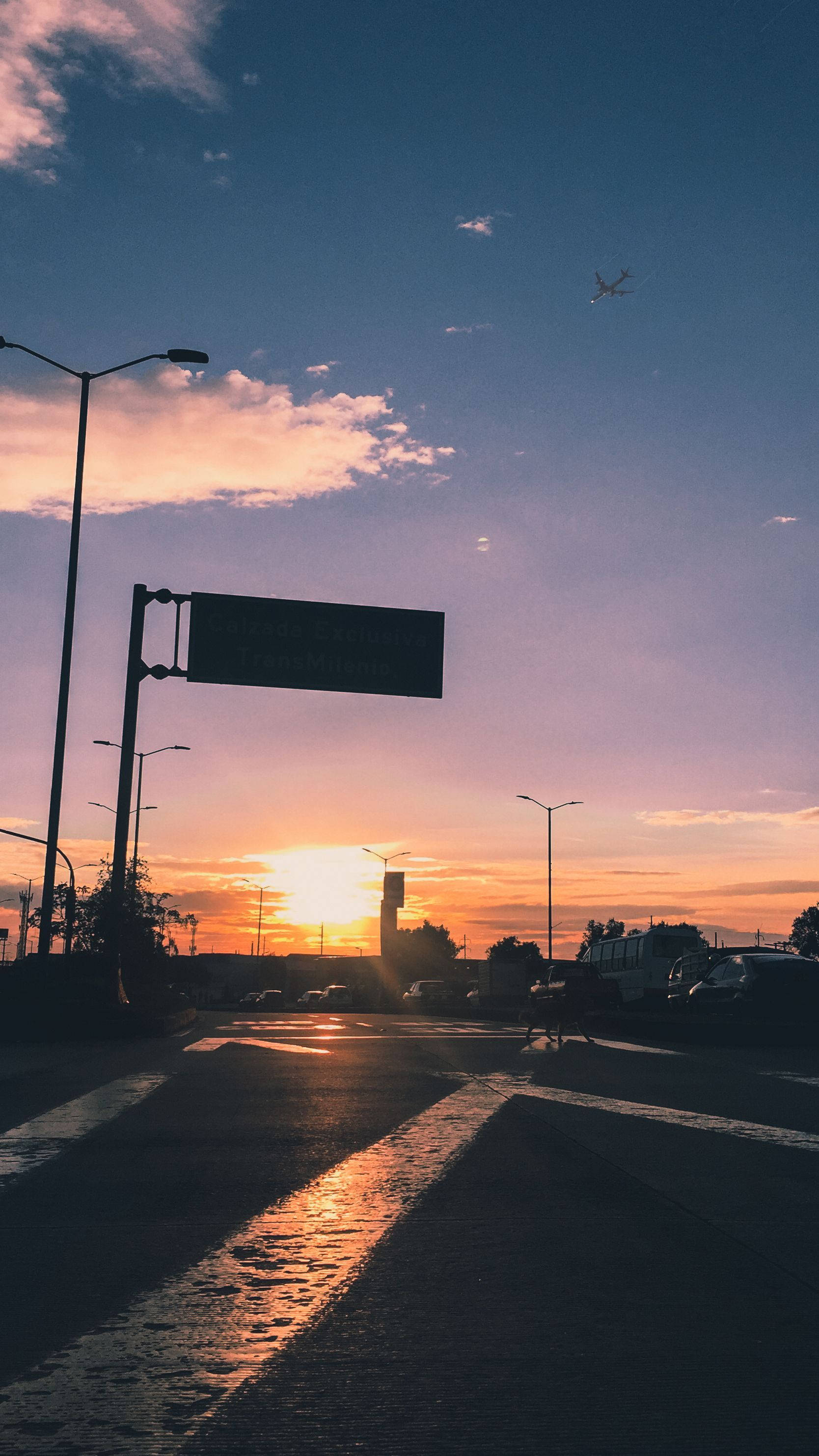 哥倫比亞波哥大 夕陽飛機 壁紙 — 夕陽將天空染成橙色和紫色，一架孤獨的飛機飛過濕漉漉的城市街道，街燈和汽車形成剪影。