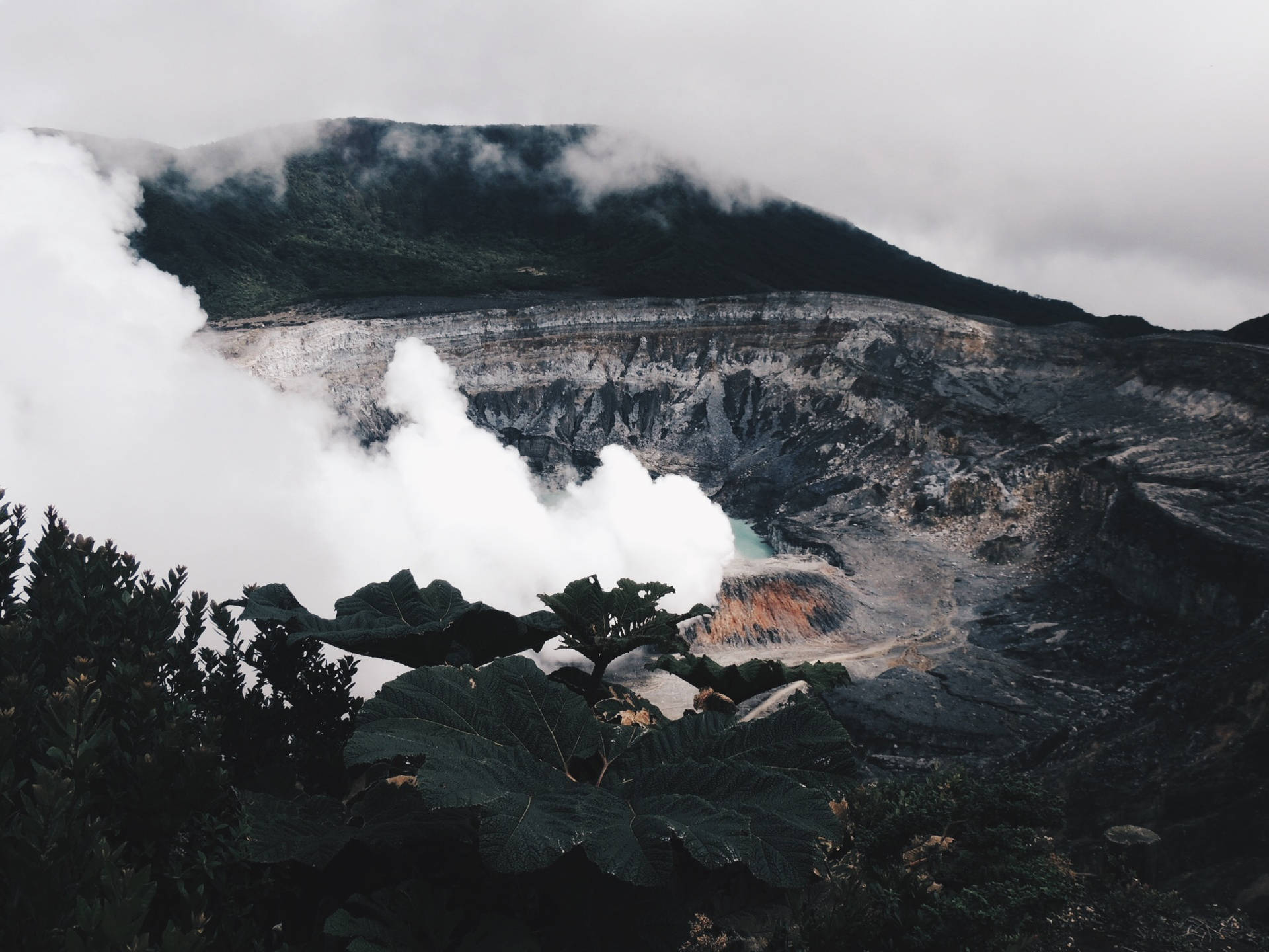 火山口噴出蒸氣，前景有藍綠色水和茂盛的綠色植物。