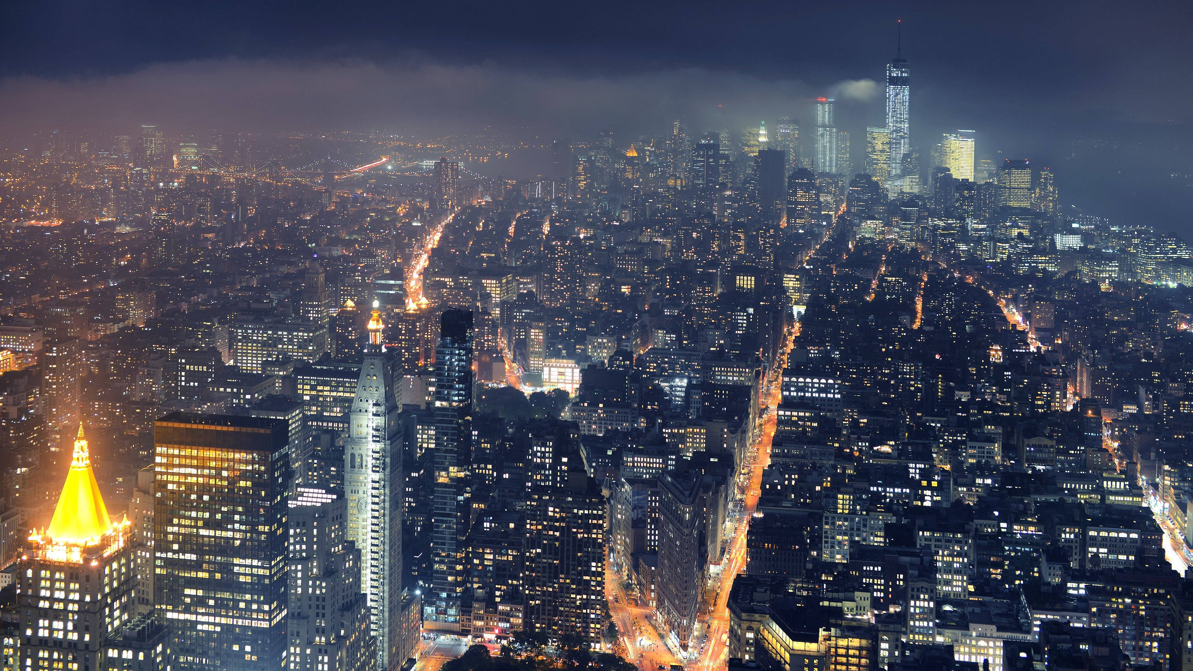 哥譚市夜景全景 壁紙 — 夜晚紐約市的鳥瞰圖，摩天大樓閃閃發光，街道燈火通明，天空陰暗多雲。