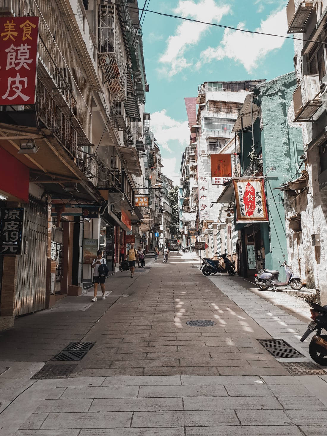 唐人街單點透視攝影 壁紙 — 一條狹窄、陽光普照的小巷，位於繁華的亞洲城市，兩旁有色彩繽紛的商店招牌和停泊的電單車。