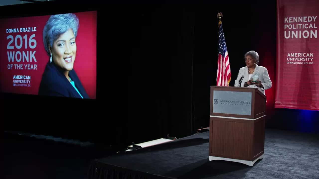 唐娜·布拉齊爾在活動中發表演講 壁紙 — Donna Brazile 站在燈光昏暗的舞台講台上，旁邊有美國國旗，身後的大屏幕顯示她的肖像以及美國大學頒發的「2016 Wonk of the Year」獎項。