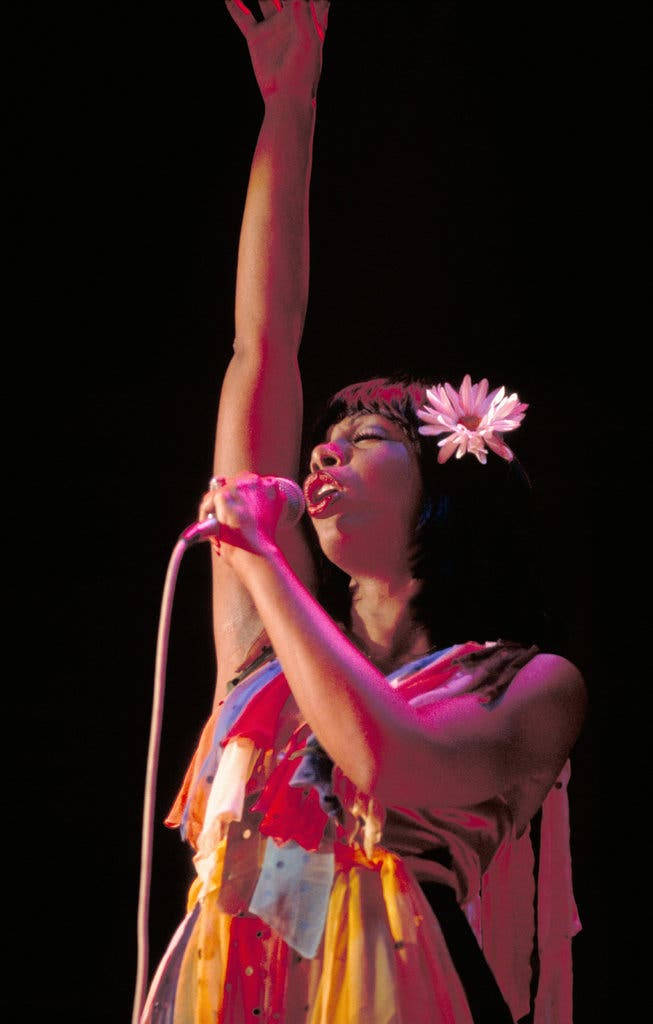 唐娜·蘇默美國現場演出 壁紙 — 一位表演者頭髮上插著粉紅色花朵，在黑色背景的舞台燈光下熱情地對著麥克風唱歌。