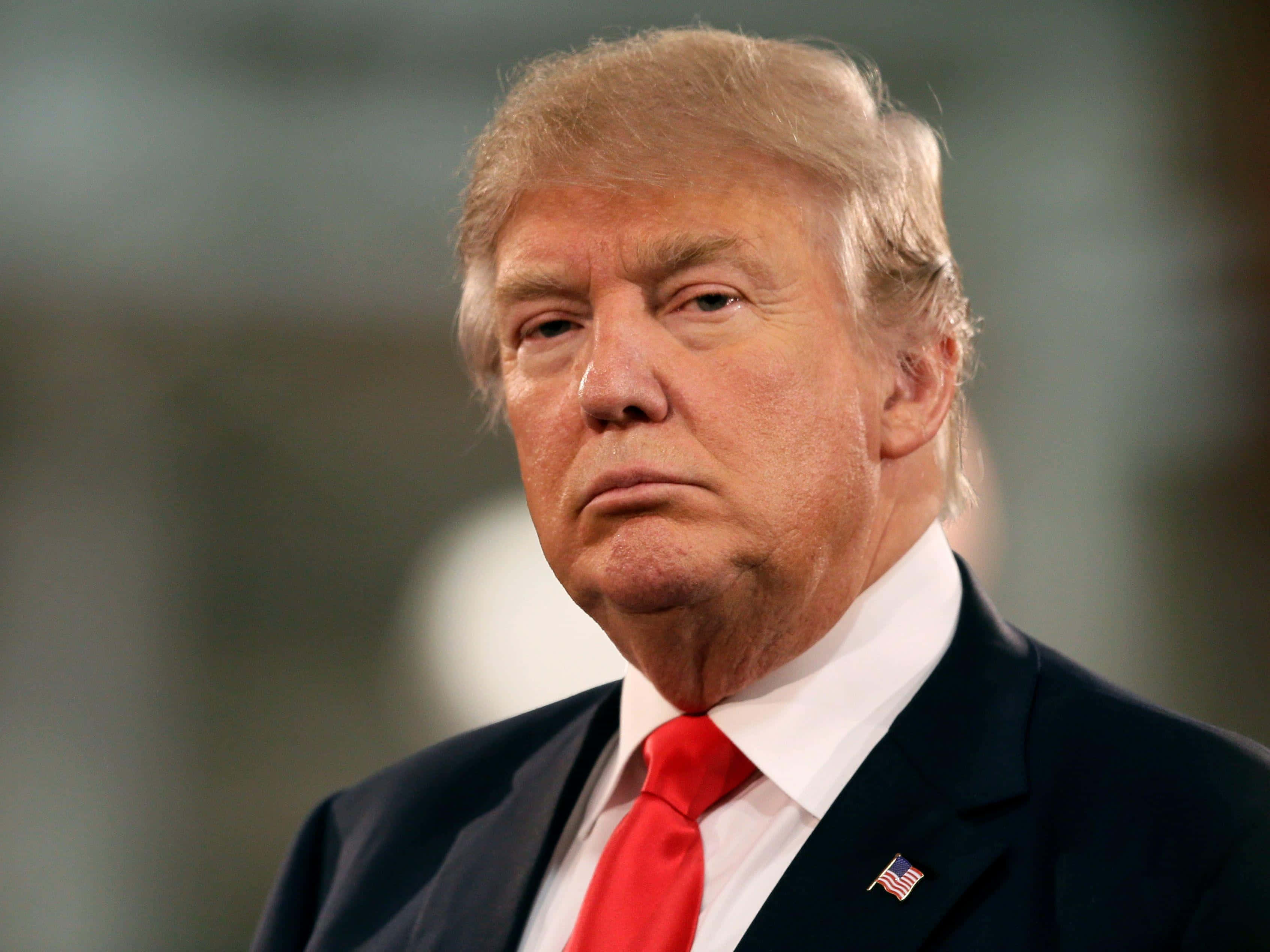 trump 桌布 壁紙 — 唐納德·特朗普嘅特寫肖像，身穿深色西裝同紅色領呔，表情嚴肅，背景模糊。