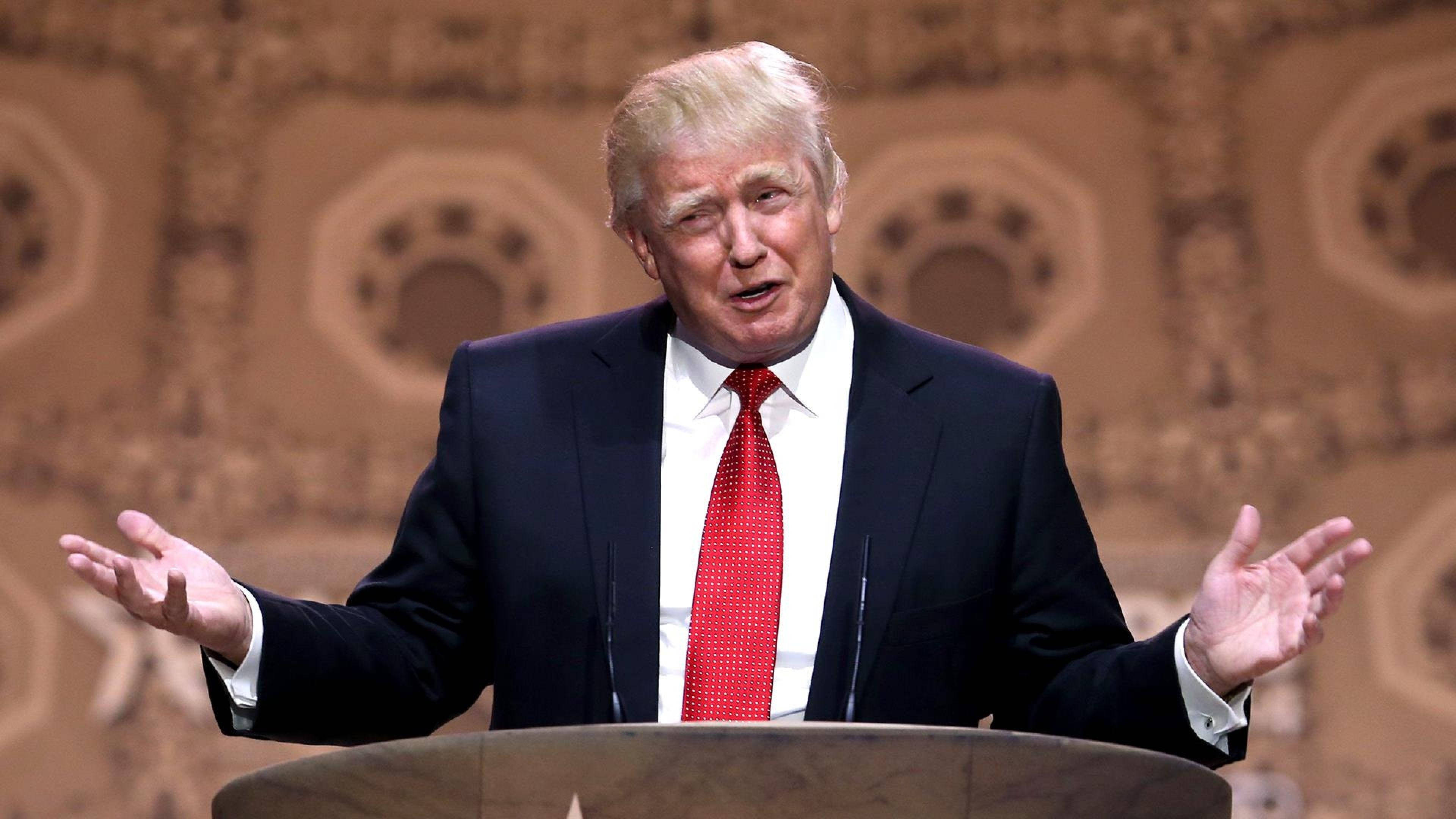 donald trump 桌布 壁紙 — 唐納德·特朗普在華麗圖案背景前，於講台上大動作地比手畫腳發言。