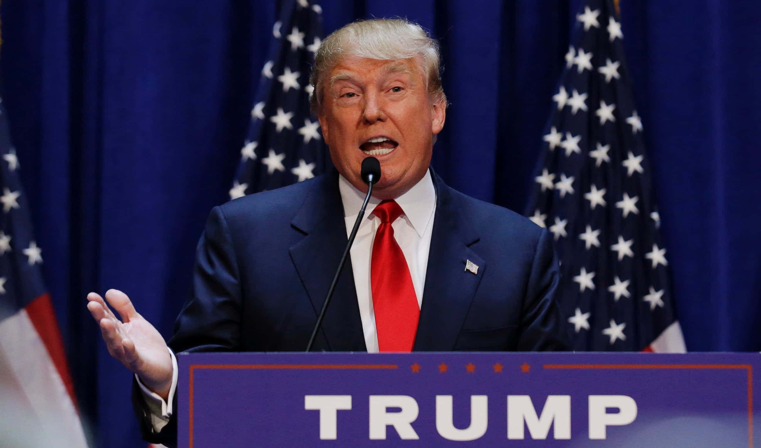 trump 桌布 壁紙 — 唐納德·特朗普在藍色帷幕前，於兩旁美國國旗簇擁下，在帶有「TRUMP」標誌的講台上慷慨激昂地發言。
