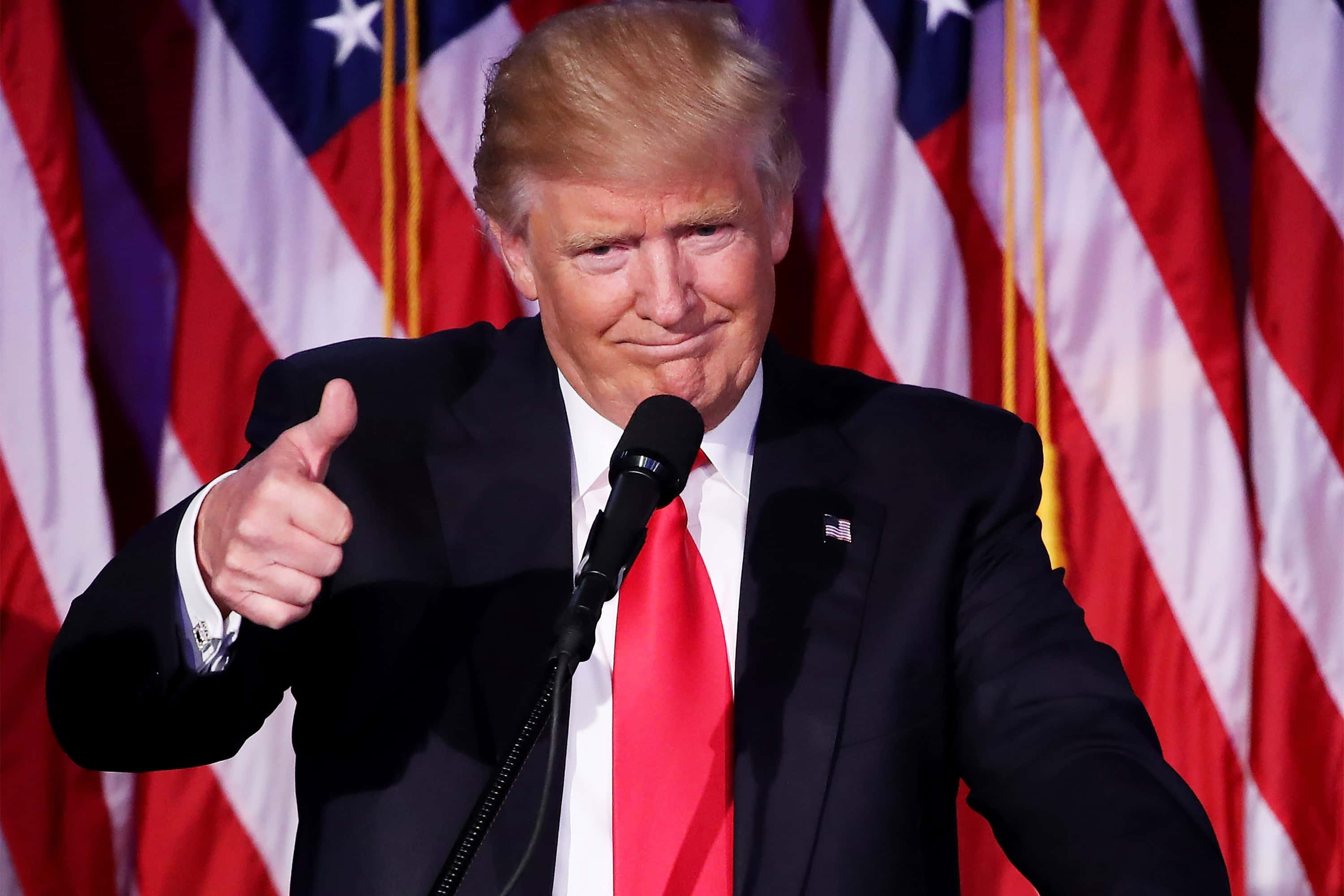 trump 桌布 壁紙 — 唐納德·特朗普在講台上發言時豎起大拇指，身後有美國國旗。