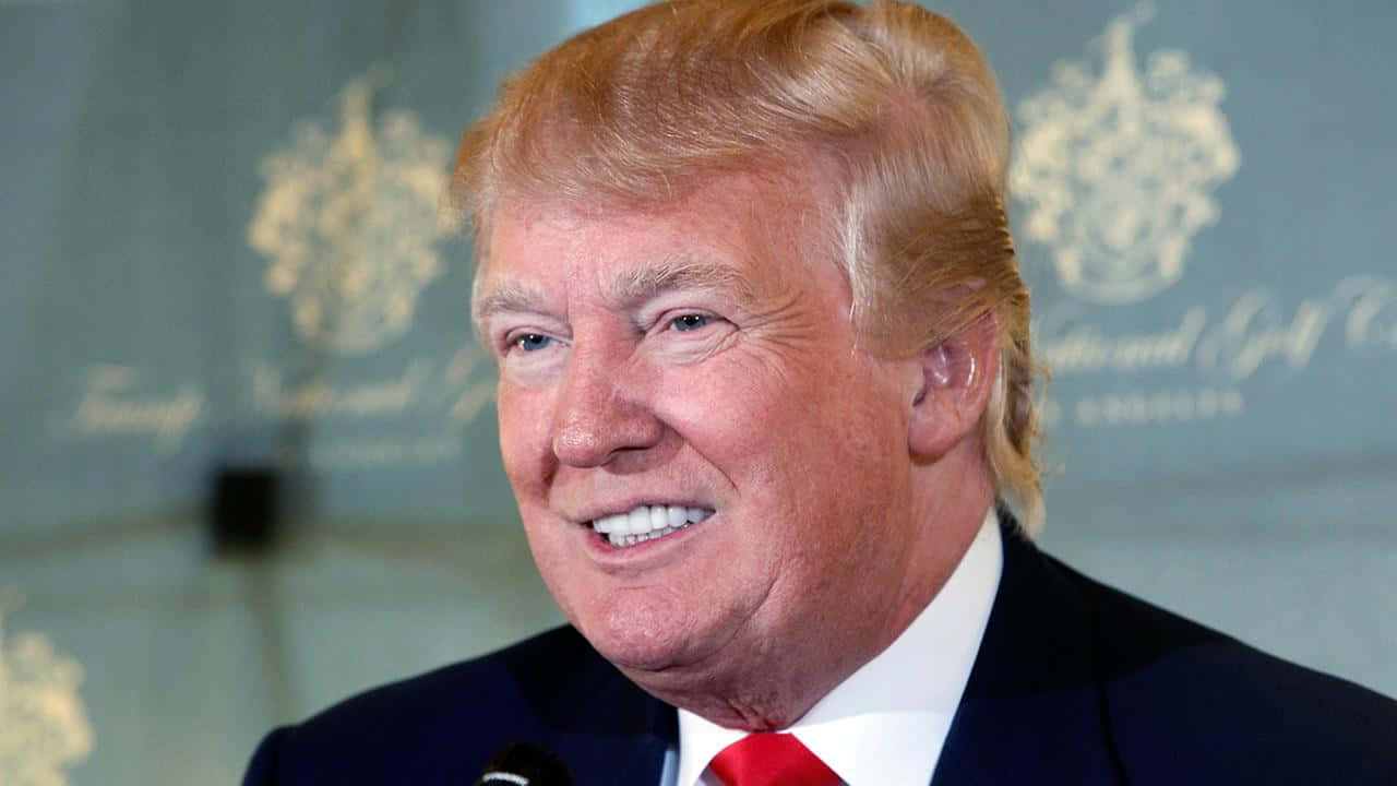 trump 桌布 壁紙 — 唐納德·特朗普的特寫，身穿深色西裝和紅色領帶，在模糊的高爾夫球場背景下微笑。