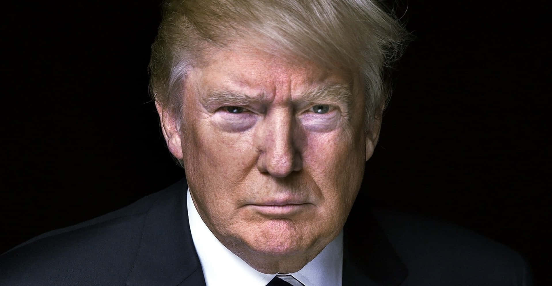 trump 桌布 壁紙 — 唐納德·特朗普身穿深色西裝，在黑色背景前的特寫肖像，帶有強烈的直視目光。