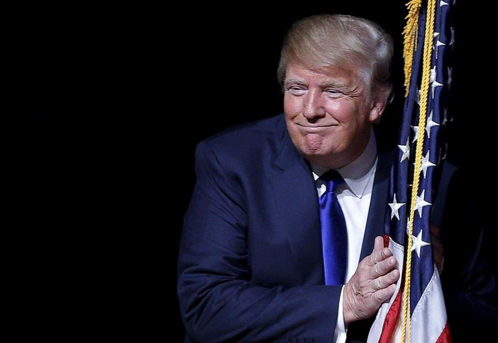 trump 桌布 壁紙 — 唐納德·特朗普身穿深色西裝，在黑色背景前微笑著手持美國國旗。