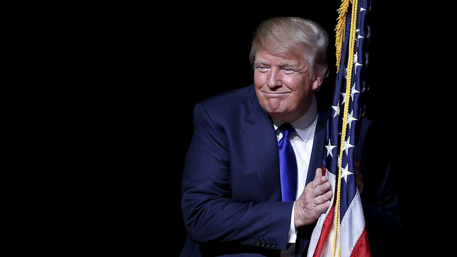 trump 桌布 壁紙 — 唐納德·特朗普身穿深色西裝，微笑著手持美國國旗，背景為黑色。