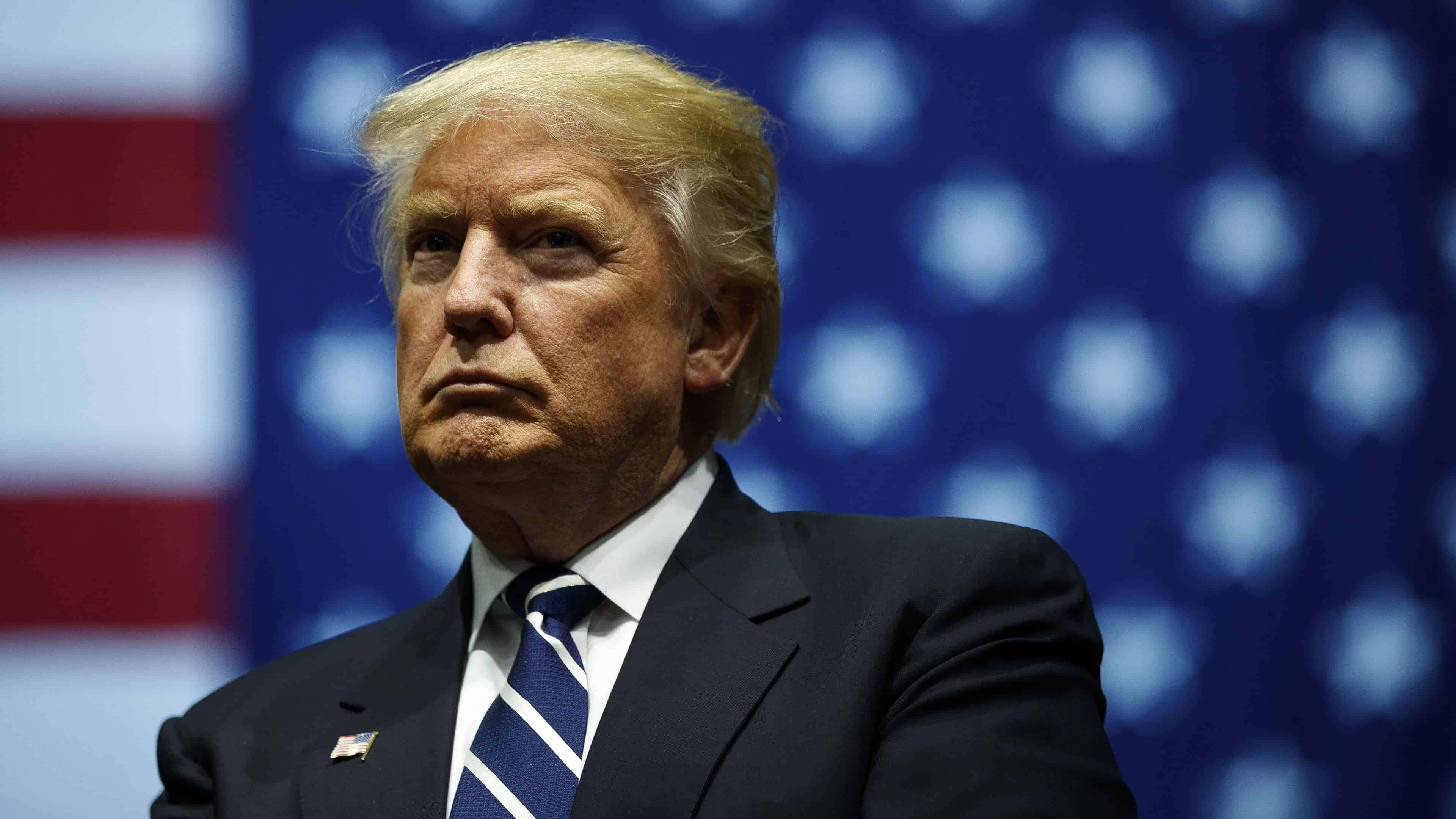 trump 桌布 壁紙 — 唐納德·特朗普身穿深色西裝和條紋領帶，莊嚴地站在模糊的美國國旗背景前。