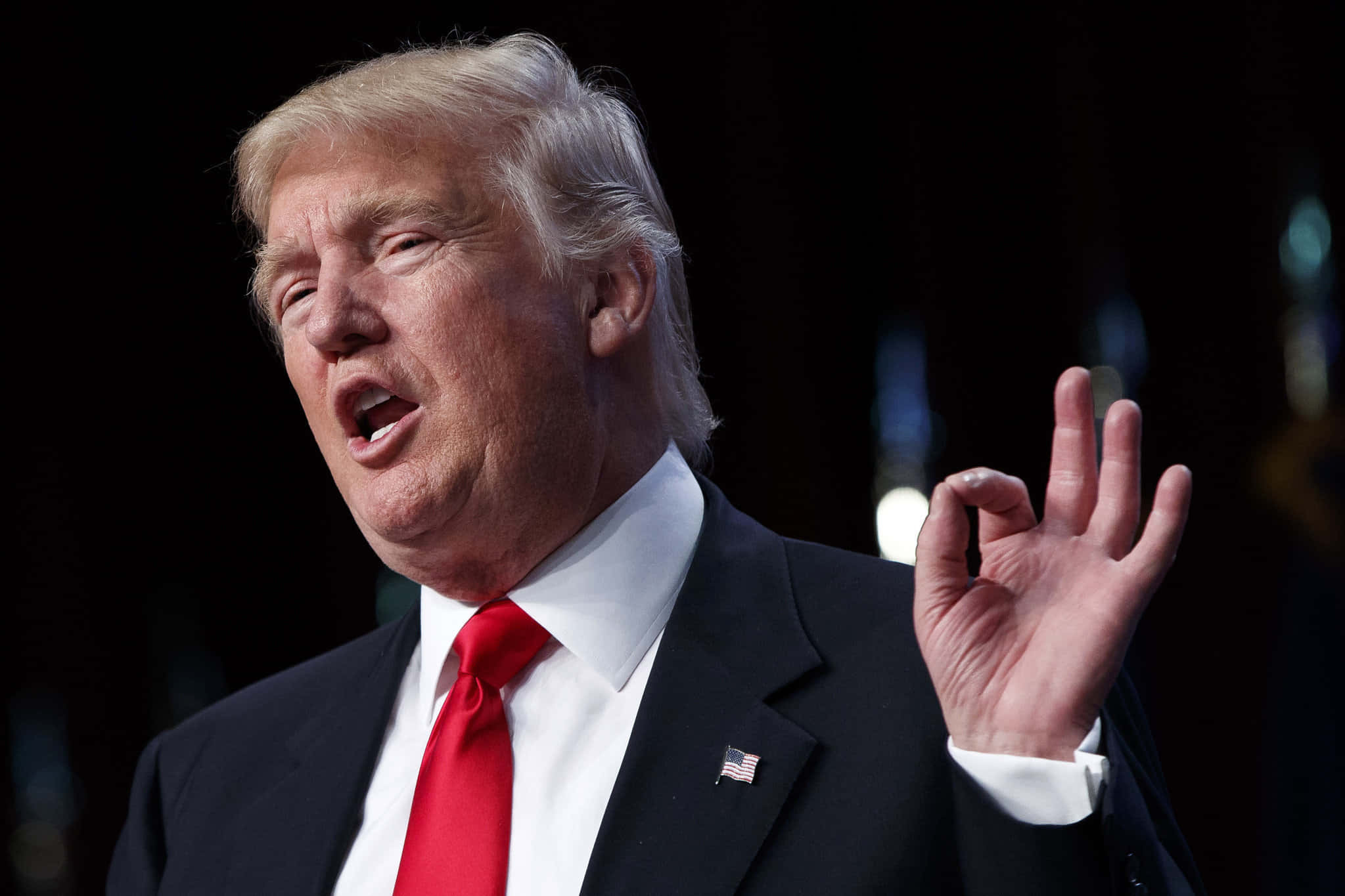 trump 桌布 壁紙 — 唐納德·特朗普身穿深色西裝和紅色領帶，在模糊的深色背景下做出「OK」手勢。