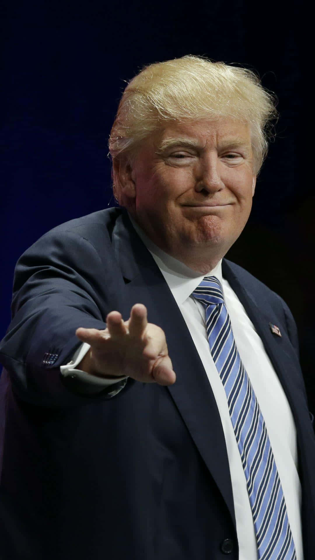 trump 桌布 壁紙 — 唐納德·特朗普身穿深色西裝和藍色條紋領帶，在深色背景前自信地微笑並向前做手勢。