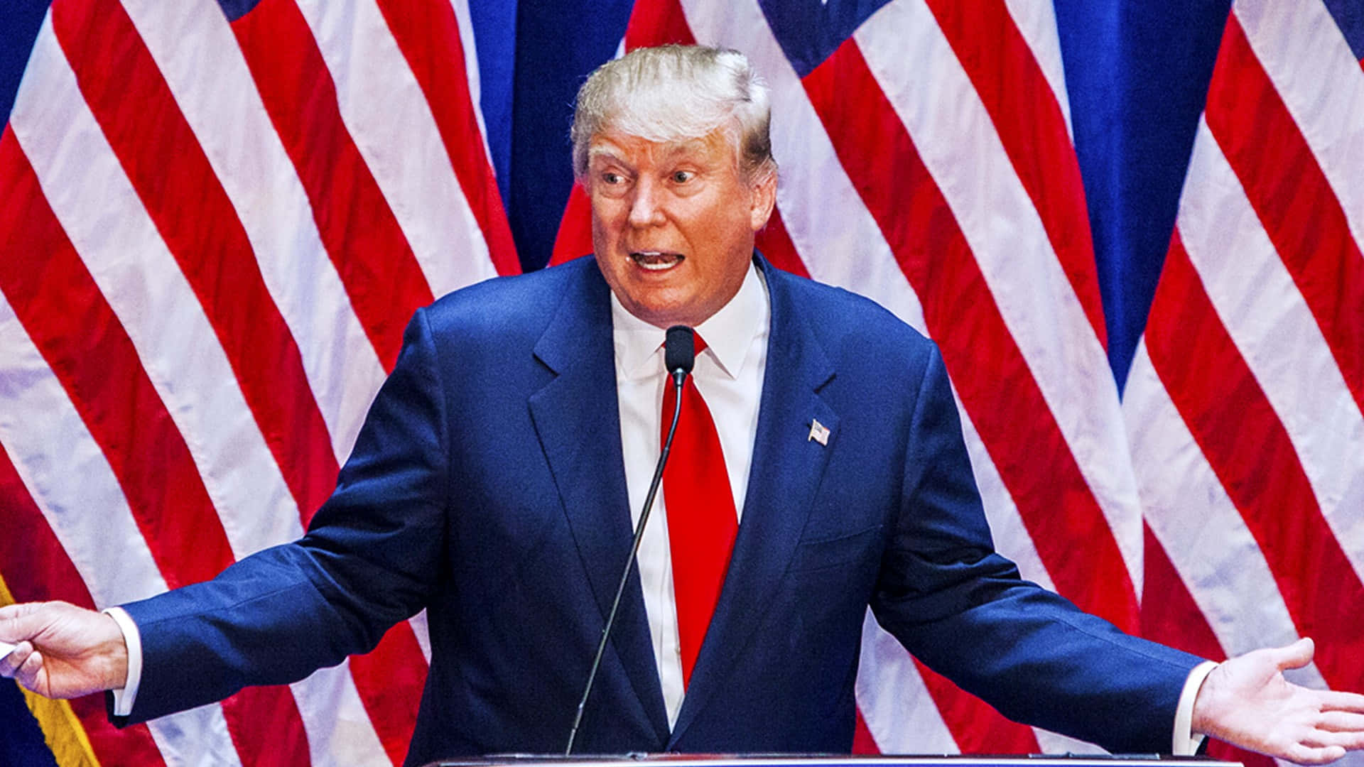 trump 桌布 壁紙 — 唐納德·特朗普身穿藍色西裝和紅色領帶，在身後有美國國旗的講台上發言。