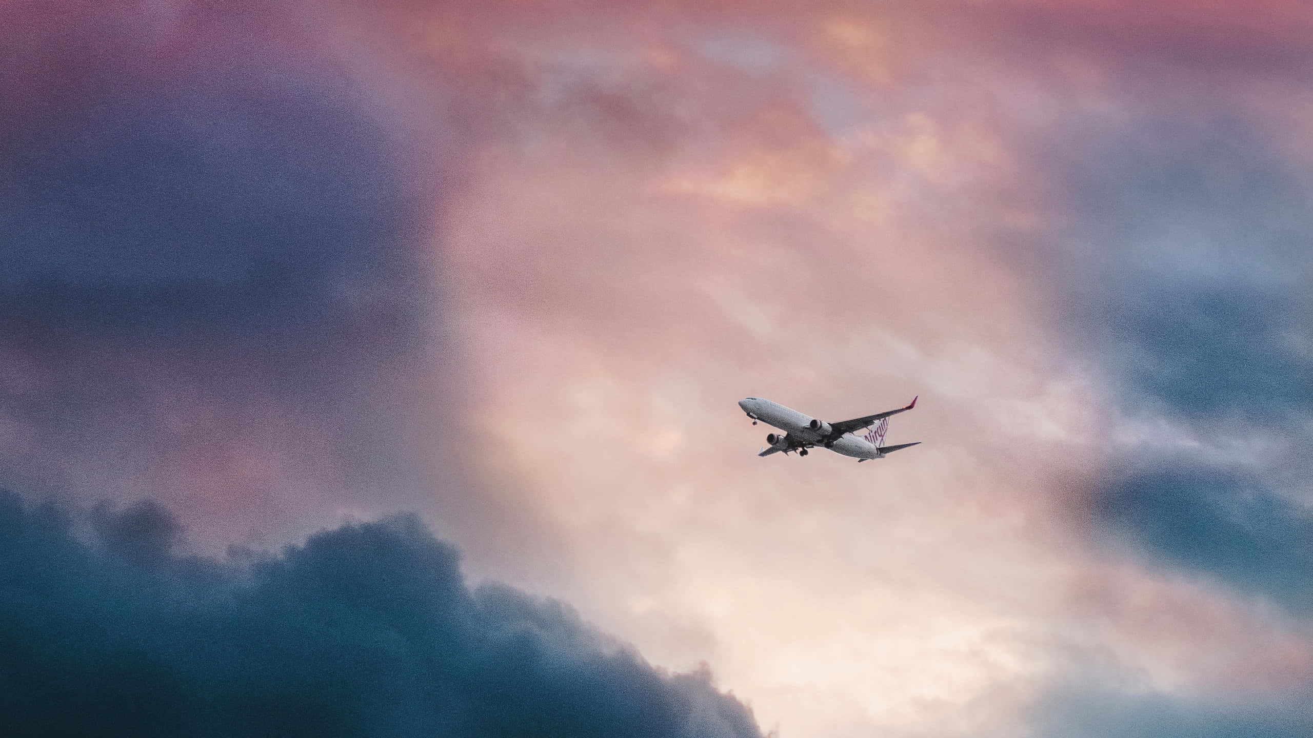 商業客機空中視角體驗 壁紙 — 一架商用飛機在暮色中翱翔於由粉紅、紫色和藍色雲彩繪成的戲劇性天空。