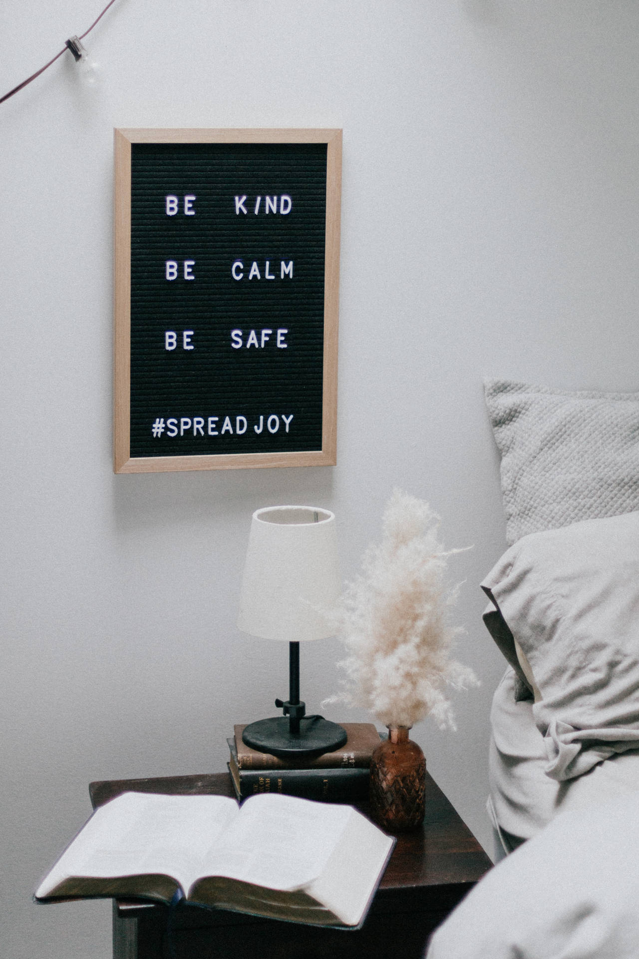 舒適的臥室角落，字母板上寫著 be kind be calm be safe spread joy，放在有燈、書和蒲葦的床頭櫃上方。