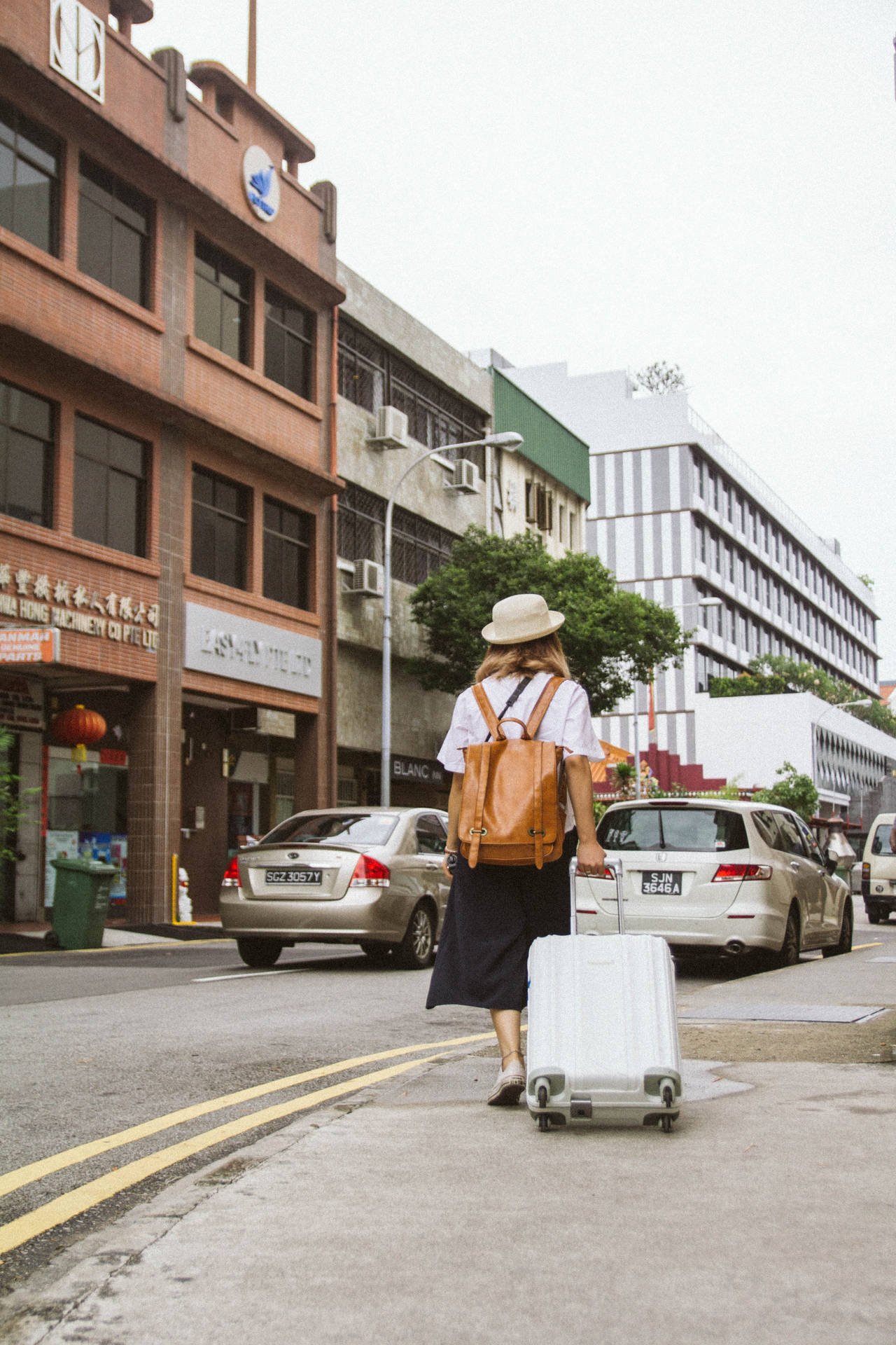 啟發獨行旅行冒險 壁紙 — 一位戴著草帽和棕色背包的旅客，拖著白色行李箱，沿著兩旁有磚砌建築和停泊汽車的城市街道行走。