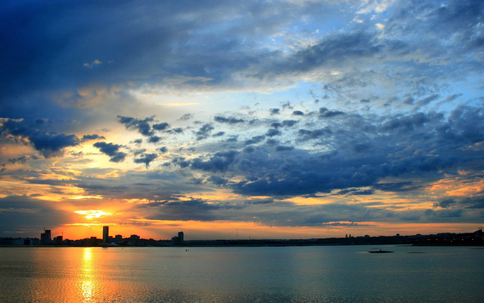 喀山城市天際線日落 壁紙 — 夕陽將天空染成火橙色和深藍色，雲朵倒映在平靜的水面上，城市天際線剪影。