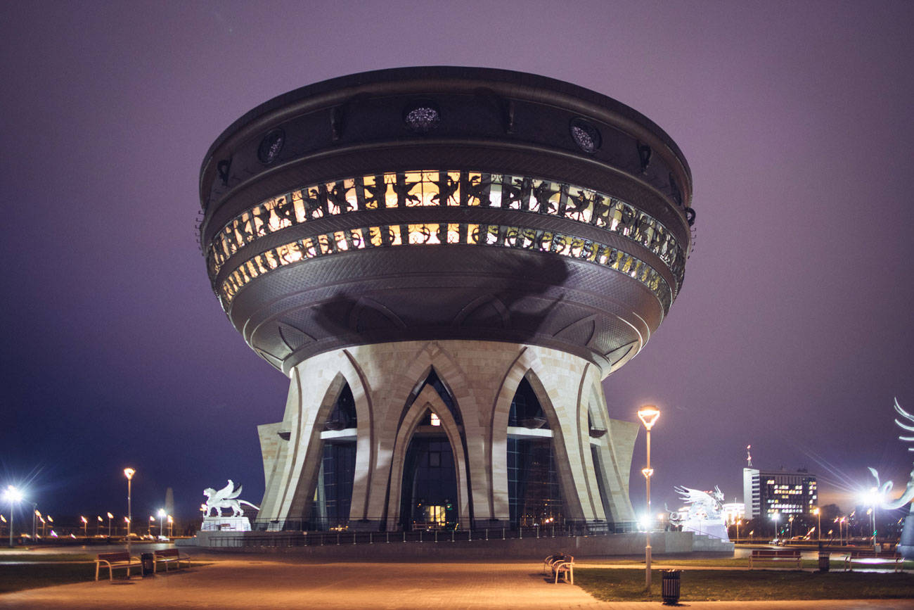 喀山婚禮宮夜景 壁紙 — 一座宏偉的圓形建築在夜晚亮起燈光，拱形支撐結構在紫色天空下散發溫暖光芒。