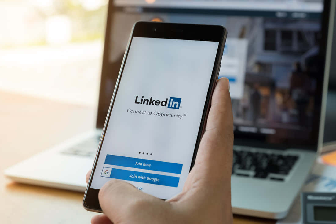 善用LinkedIn專業社交網絡的力量 壁紙 — 一隻手拿著智能手機，顯示LinkedIn登入畫面，背景有一部手提電腦。