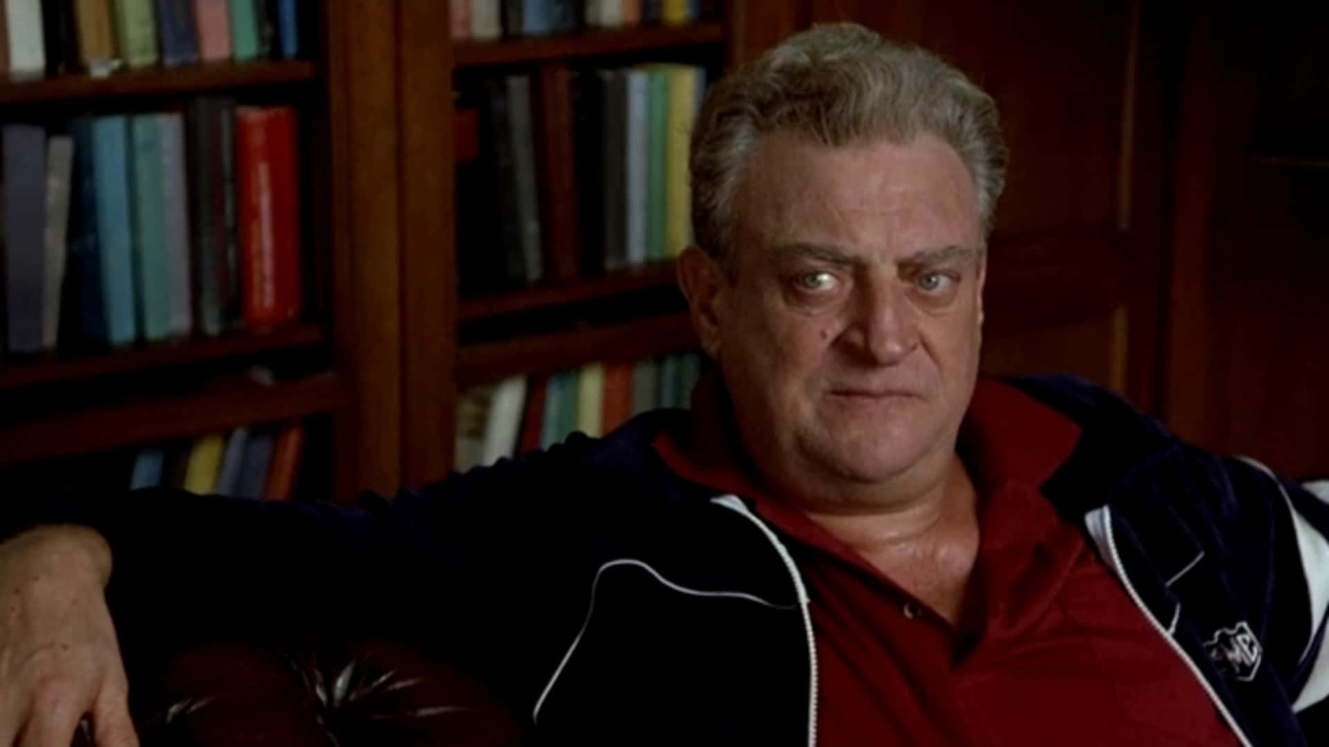 一位身穿紅襯衫和黑色外套的男人坐在皮椅上，身後有書架。