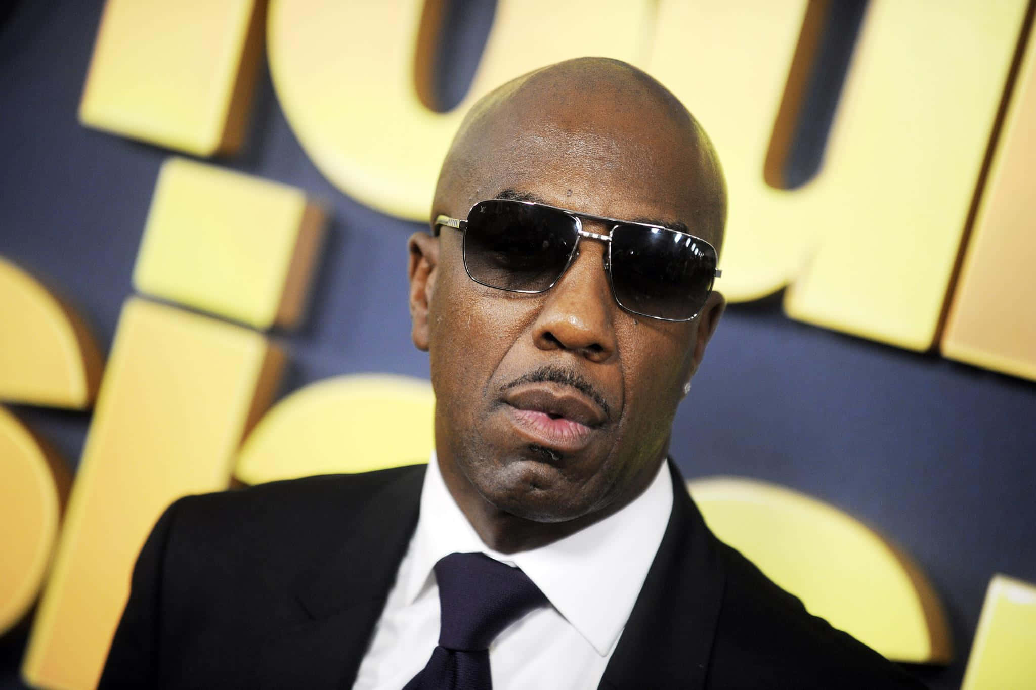 喜劇演員兼演員 J.B. Smoove 的肖像照 壁紙 — 一名身穿黑色西裝和太陽眼鏡的男子在鮮明的黃色和藍色背景前自信地擺姿勢。