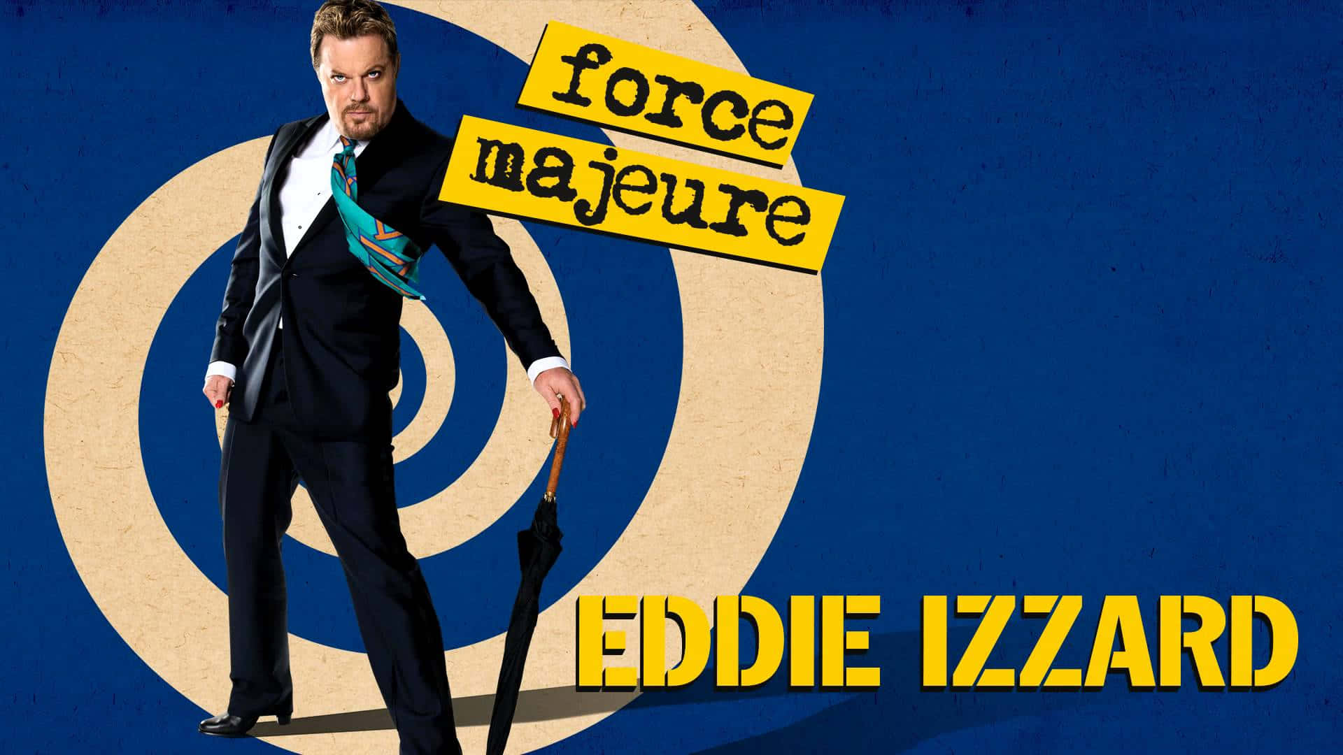 Eddie Izzard 桌布 壁紙 — 喜劇演員艾迪·伊扎德身穿利落西裝，自信地站在大膽的藍色背景前，背景有同心圓和黃色文字「force majeure」。