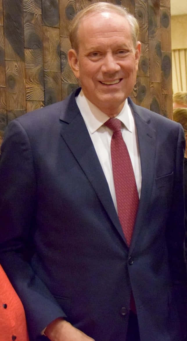 一位身穿深藍色西裝和紅色領帶的男子在室內對著木牆微笑。