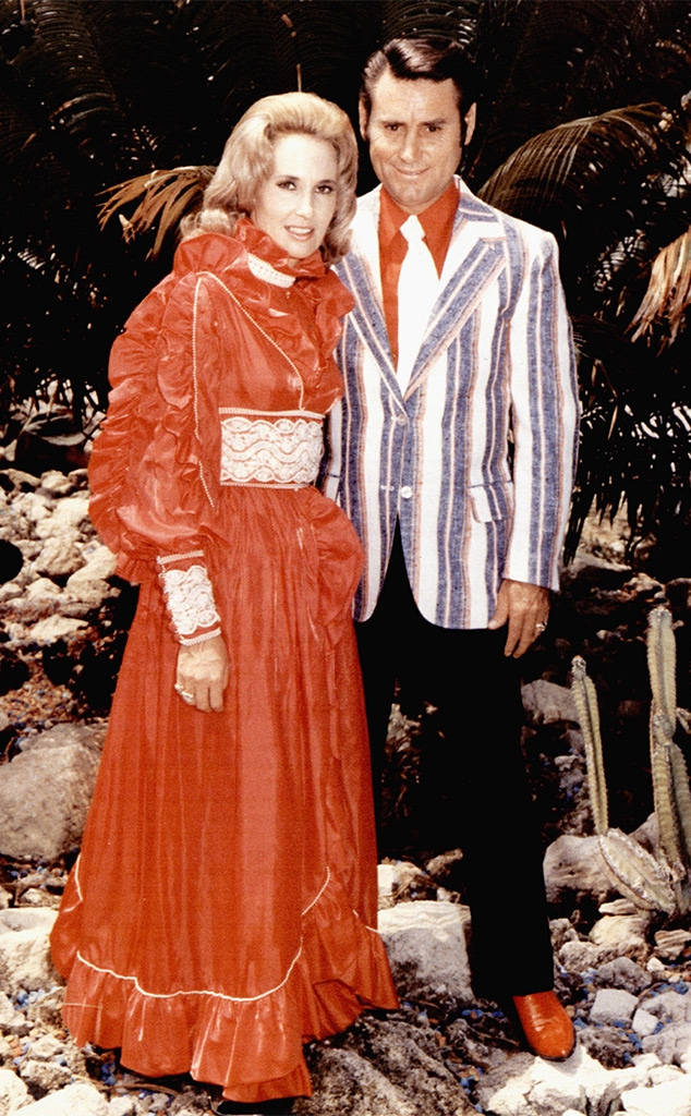 喬治·瓊斯與塔米·溫內特——傳奇鄉村音樂情侶 壁紙 — 一對魅力四射的情侶穿著大膽的1970年代服裝，在棕櫚葉點綴的岩石熱帶背景下自信地擺姿勢。
