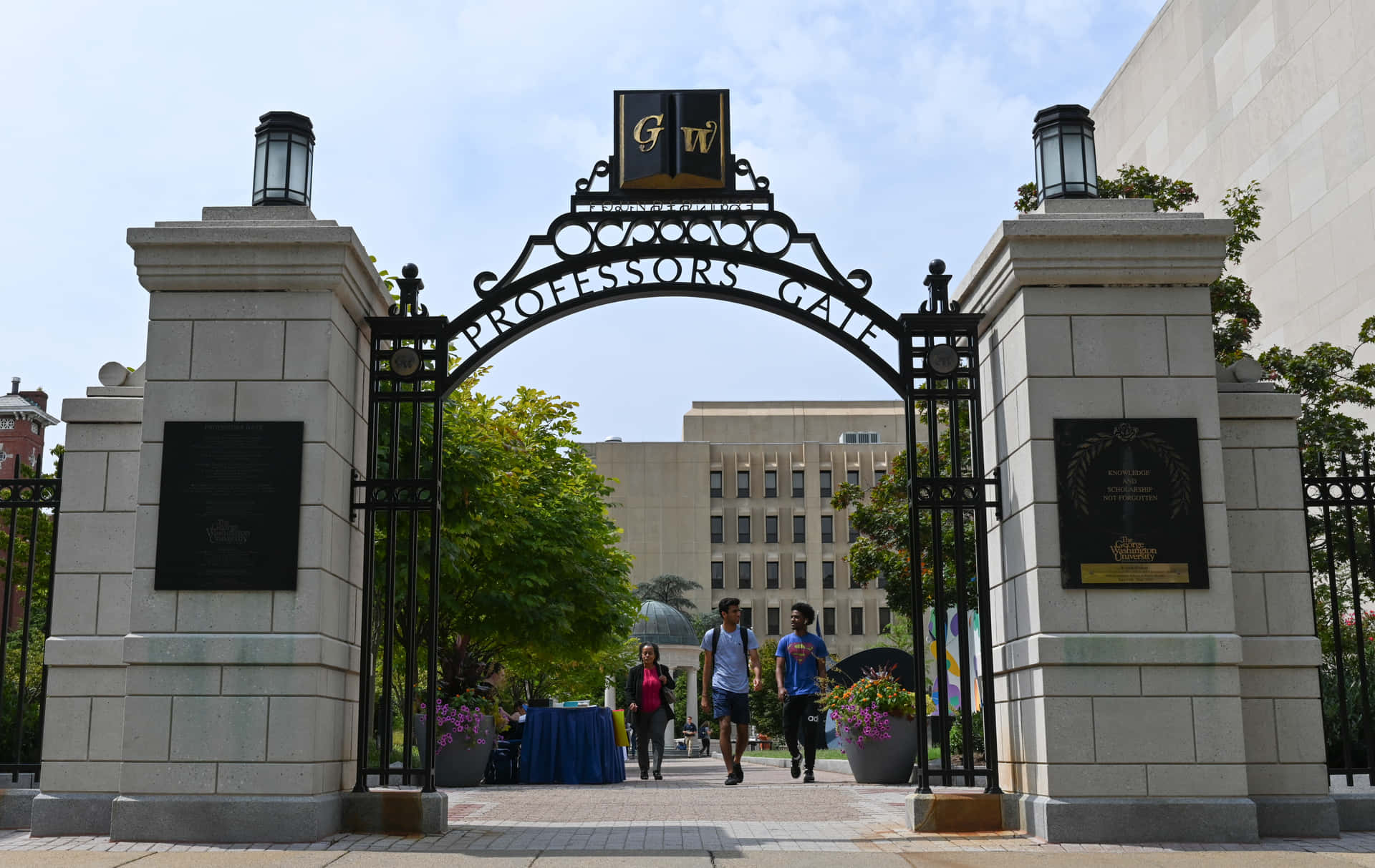 人們在陽光明媚的日子裡穿過寫有 Professors Gate 的大學大門。