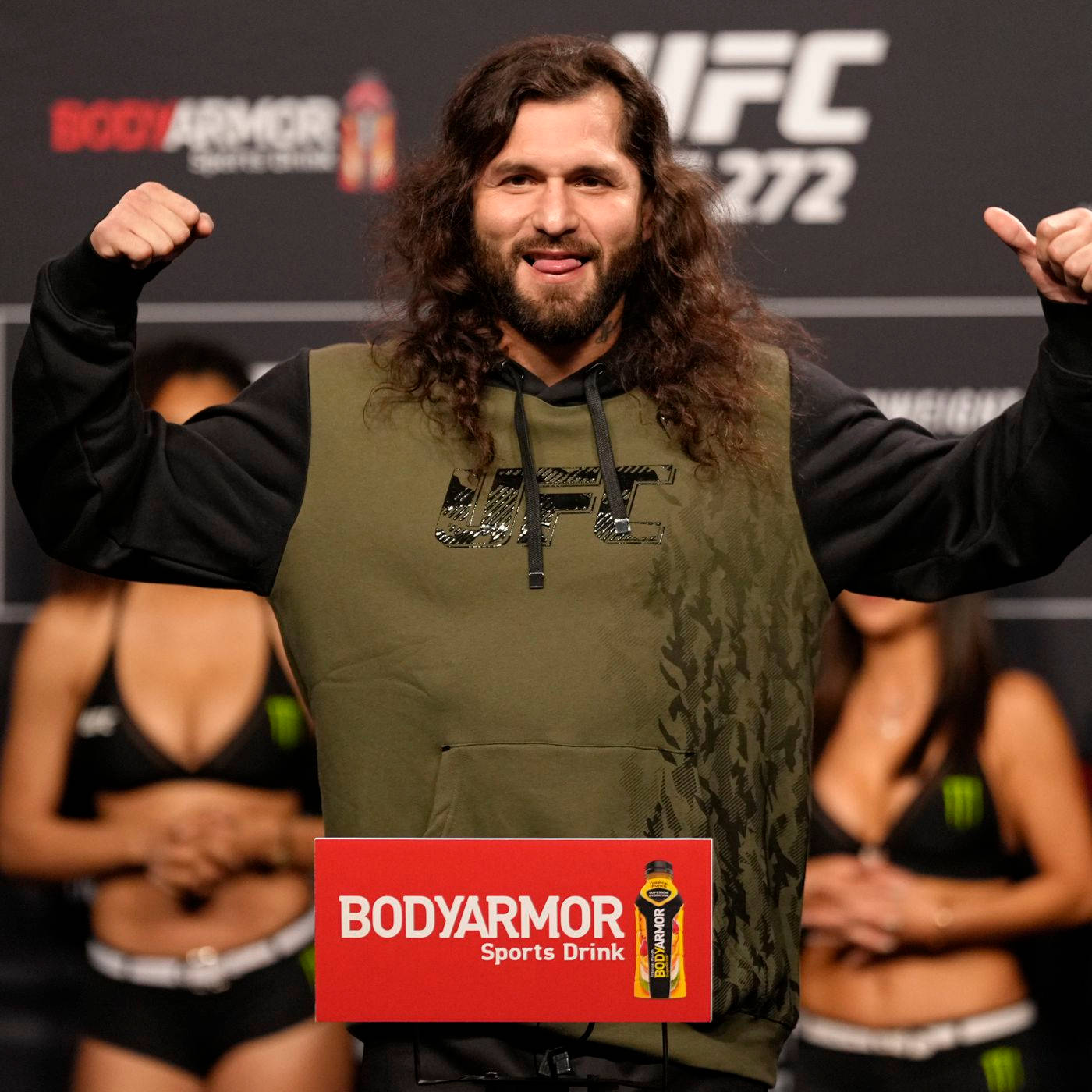 喬爾格·馬斯維達爾穿連帽衫 壁紙 — 一位留著長捲髮、留鬍子的UFC拳手，穿著綠色UFC連帽衫，在稱重活動中自信地擺姿勢。