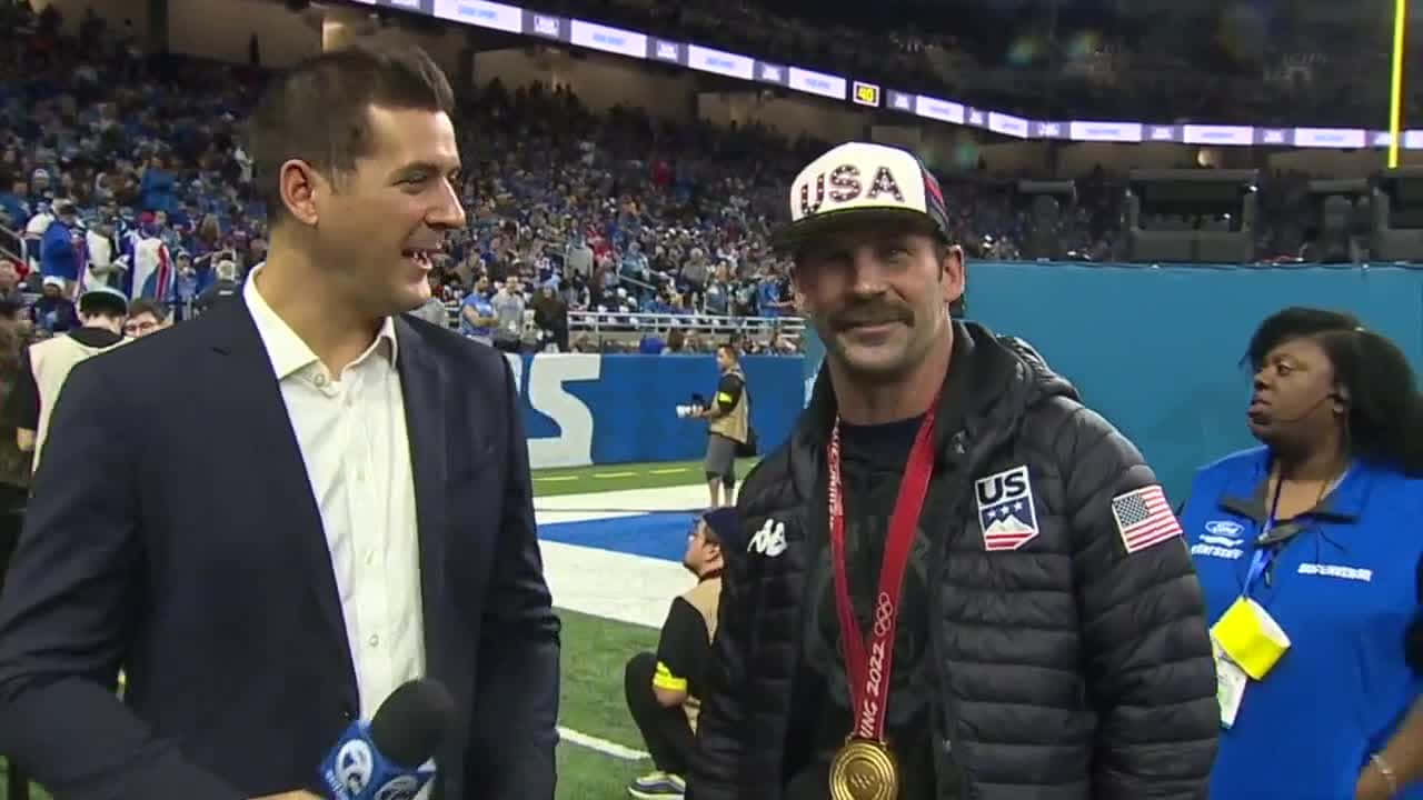 單板滑雪選手Nick Baumgartner訪問照 壁紙 — 一位面帶微笑的奧運獎牌得主，戴著美國帽和金牌，在足球場上接受記者採訪，記者手持麥克風。