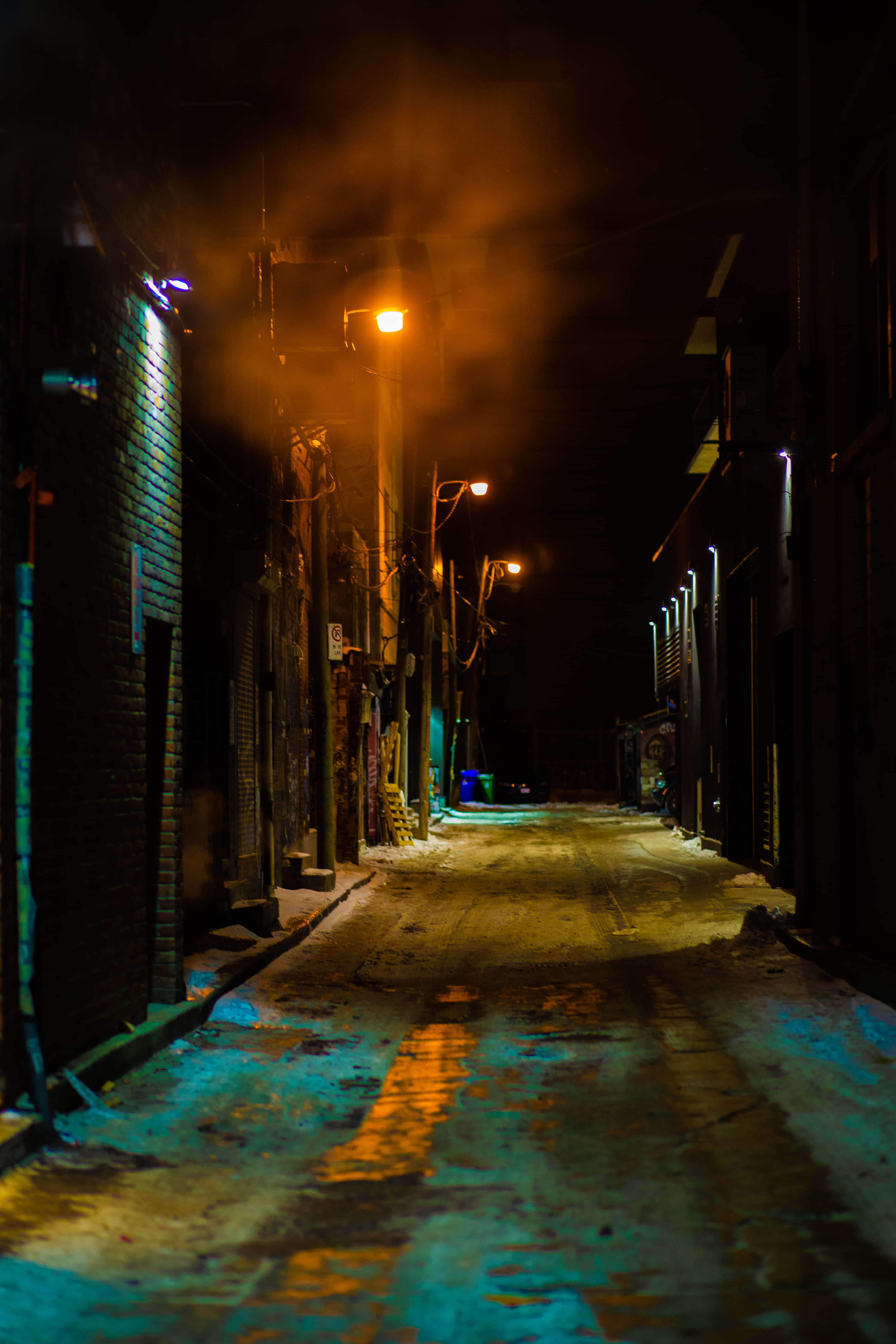 單盞街燈巷弄背景 壁紙 — 夜晚一條狹窄的積雪小巷，溫暖的路燈在潮濕的路面上投射出橙色光芒，並帶有藍色霓虹反射。